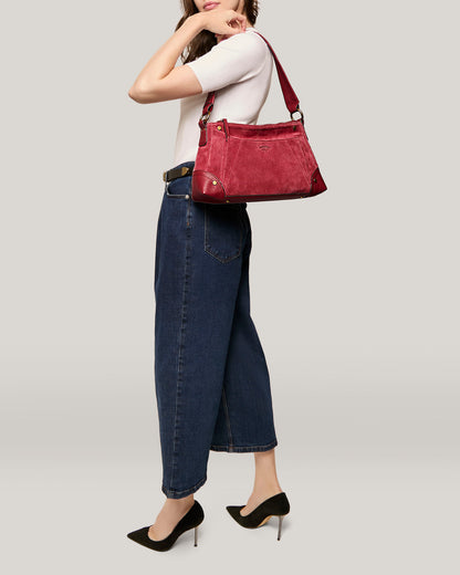 A woman is posing with the Becket Triple Entry Shoulder from American Leather Co. slung over her shoulder. She is dressed in a white top, high-waisted blue jeans, and black high-heeled shoes. Her left hand is touching her face, and the background is plain light grey.