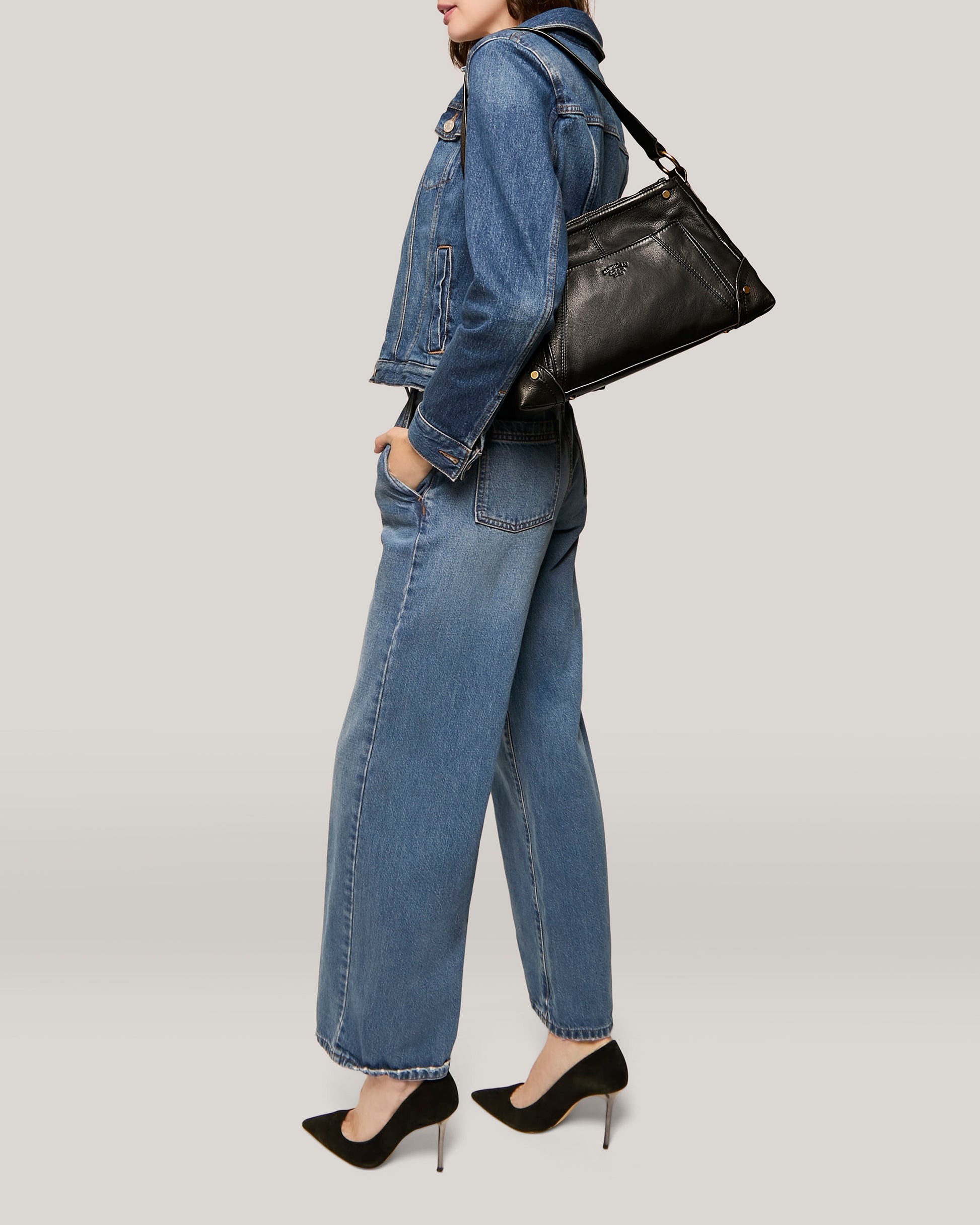 A person is wearing a denim jacket and denim wide-leg pants, accessorized with the Becket Triple Entry Shoulder bag by American Leather Co. over their shoulder and black high-heeled shoes. The background is a light, solid color. The image captures the person from the knees up, with their hands in their pockets.