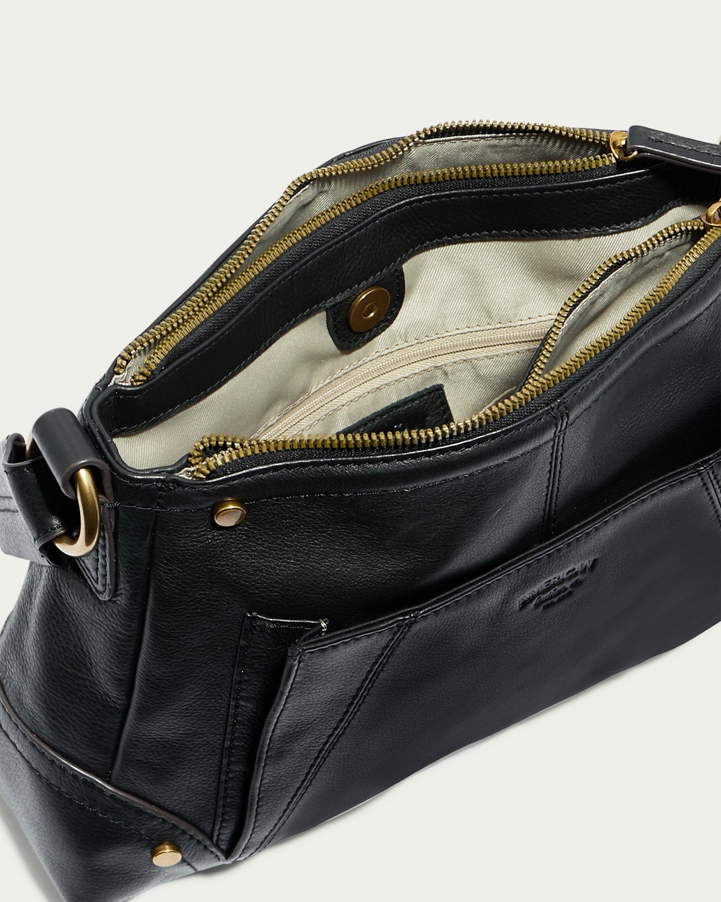 The Becket Triple Entry Shoulder Bag by American Leather Co. is an open black genuine leather shoulder bag with brass hardware, featuring spacious interior compartments. The inside boasts a beige lining, a zippered pocket, and smaller pockets for organization. This traditional shoulder bag also includes a magnetic clasp for closure and an adjustable strap.