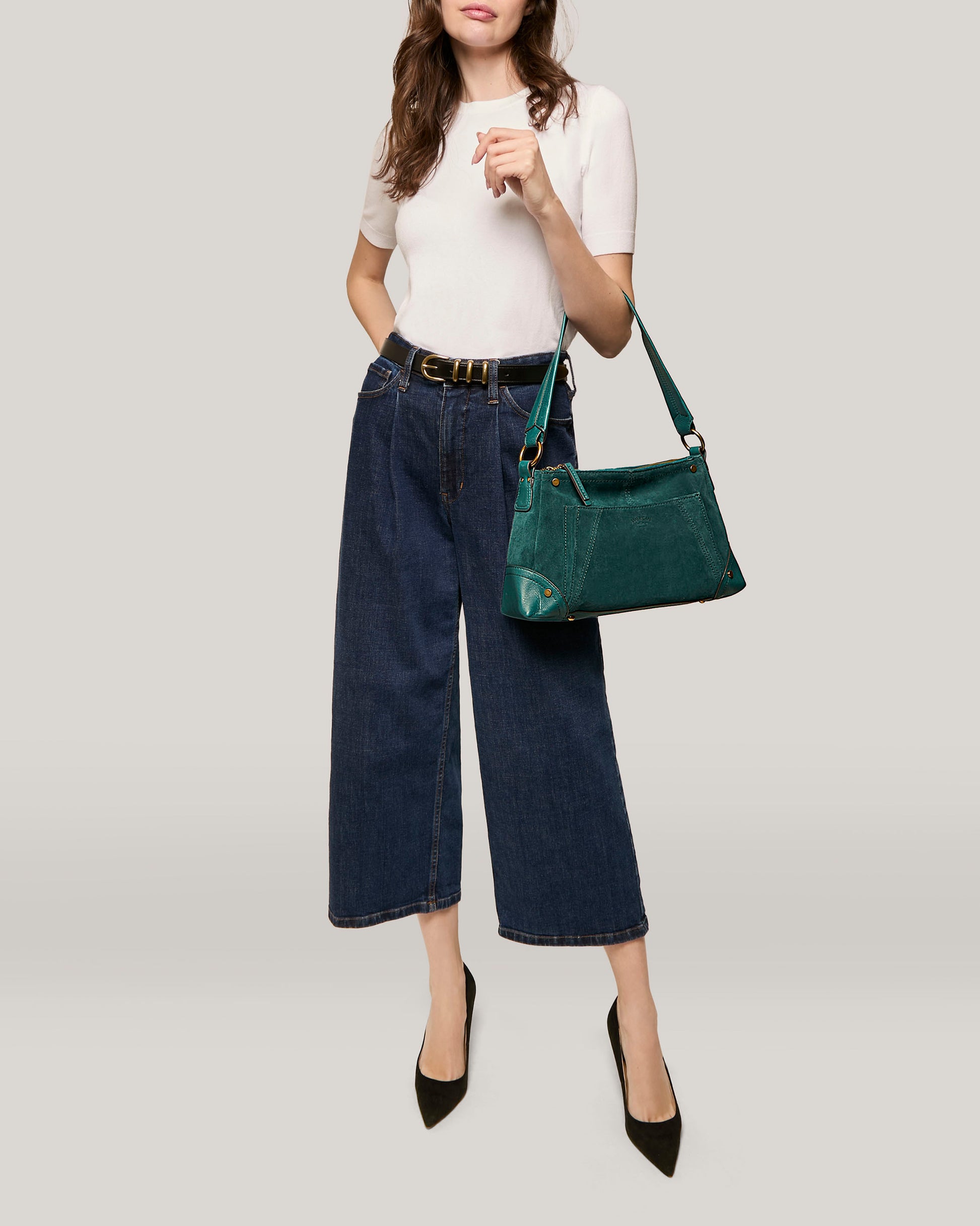 A person with long brown hair is wearing a white short-sleeve top, dark blue wide-leg cropped jeans, and black pointed high heels. They are holding an American Leather Co. Becket Triple Entry Shoulder bag made of genuine suede leather over their shoulder.