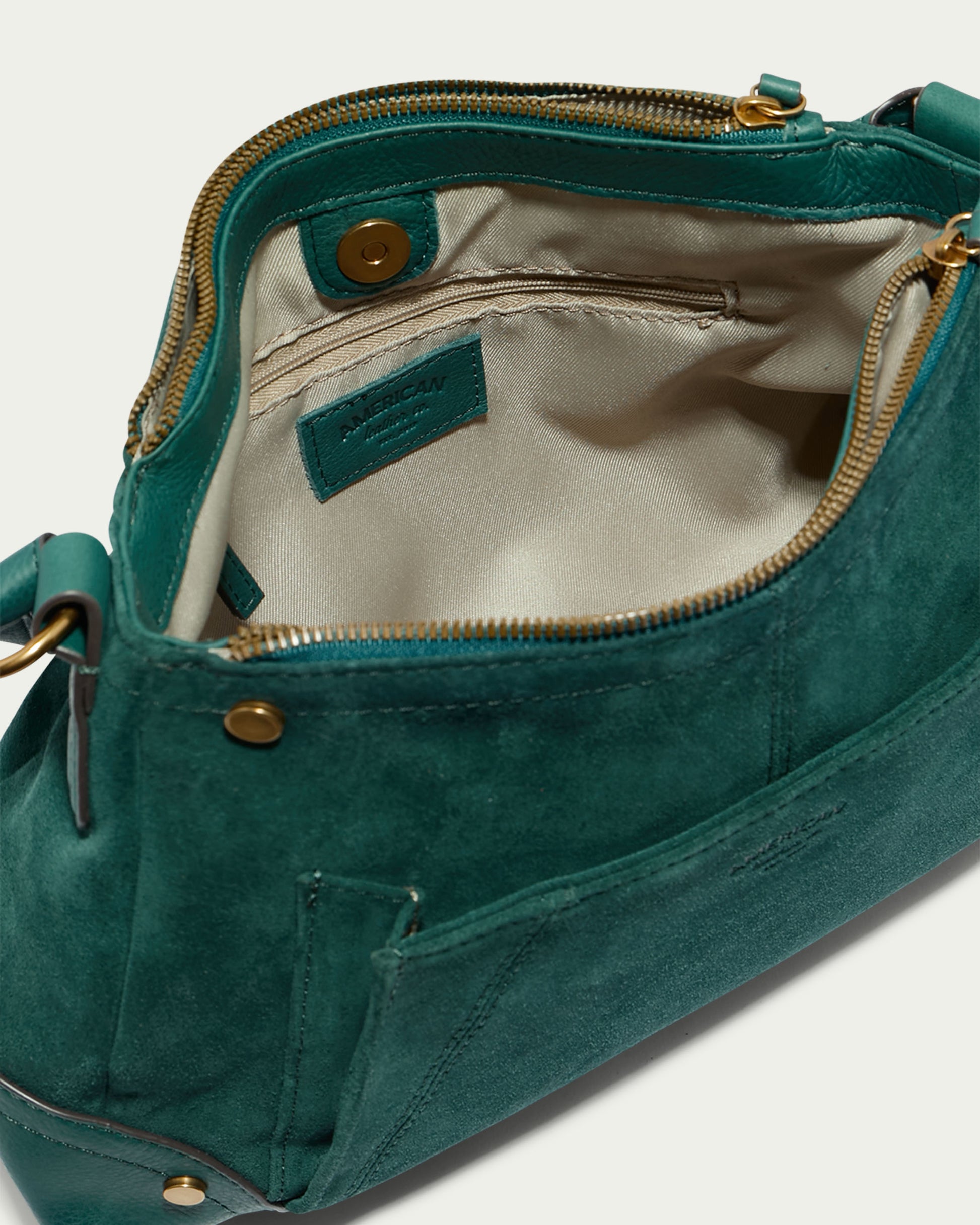 The Becket Triple Entry Shoulder handbag by American Leather Co. is a stylish green accessory made of genuine suede leather, open to reveal a beige interior lining with a label and a snap button pocket. The top edge features a gold zipper, while the exterior showcases two small rivet details. This traditional shoulder bag is angled to display its interior clearly.