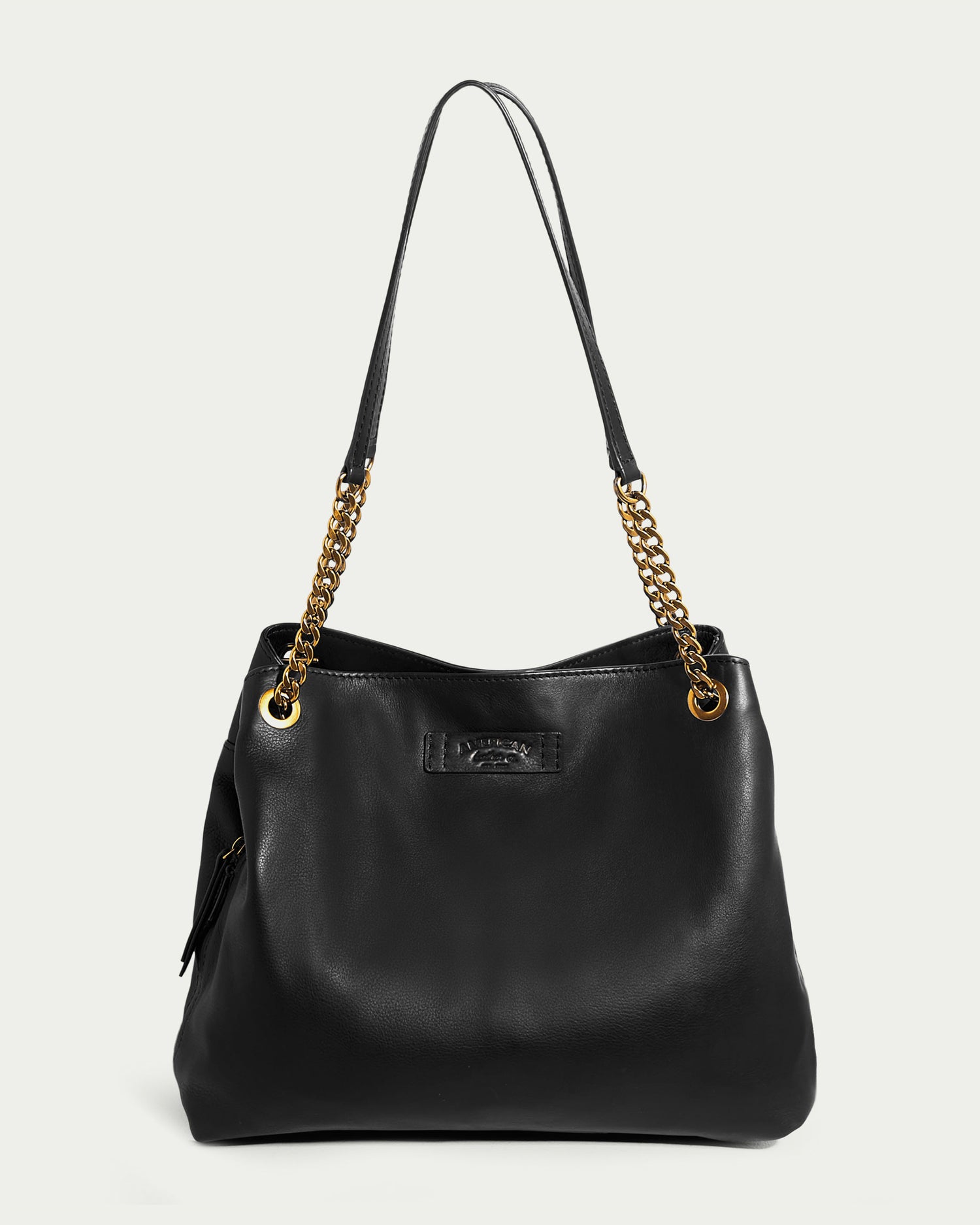 The Brisben Chain Shopper by American Leather Co. is a stylish black leather handbag with gold chain handles. It features a subtle embossed logo on the front and a side zipper pocket. Crafted from genuine leather, its sleek design and metallic accents give it a contemporary and elegant look, making it the perfect triple entry shopper.