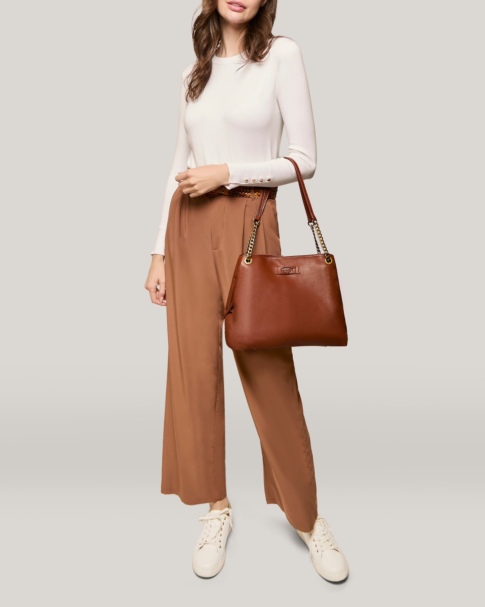 A woman dressed in a white long-sleeve top and high-waisted brown wide-leg pants stands against a plain background. She is carrying the Brisben Chain Shopper from American Leather Co. and wearing white sneakers. Her hair is loose, and she has a relaxed, confident posture with one hand resting by her side.