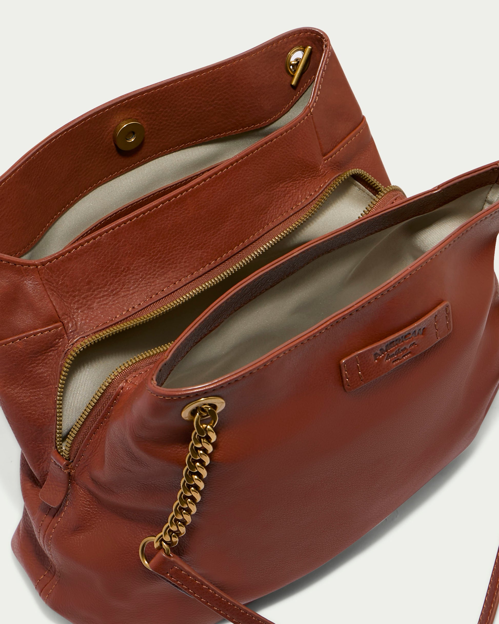 The Brisben Chain Shopper by American Leather Co., featuring a brown leather exterior and a lightweight chain handle, is shown open to reveal its interior compartments. The inside showcases a beige lining, a zippered pocket, and a magnetic snap closure for the main compartment. A leather tag with branding is also visible on the inner pocket.