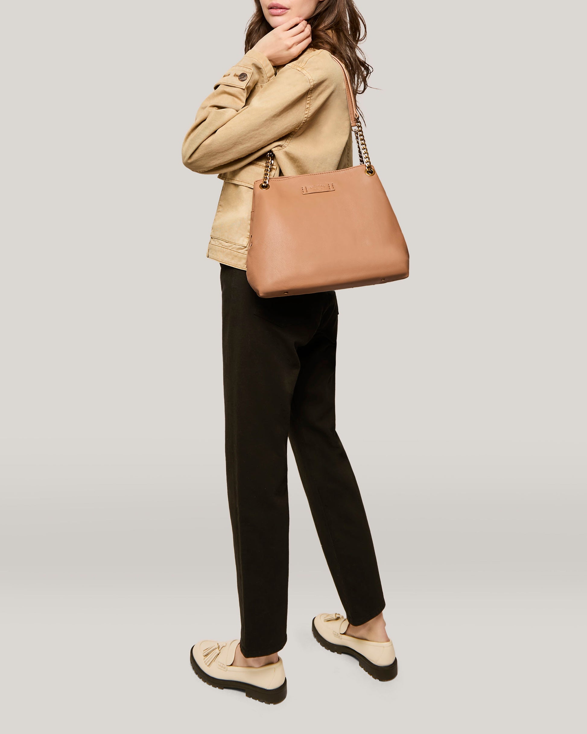 A person with long brown hair is dressed in a beige jacket and black pants. They carry an American Leather Co. Brisben Chain Shopper, a genuine leather bag with a gold chain strap, over their shoulder. They are standing against a light grey background and wearing beige loafers with black soles.