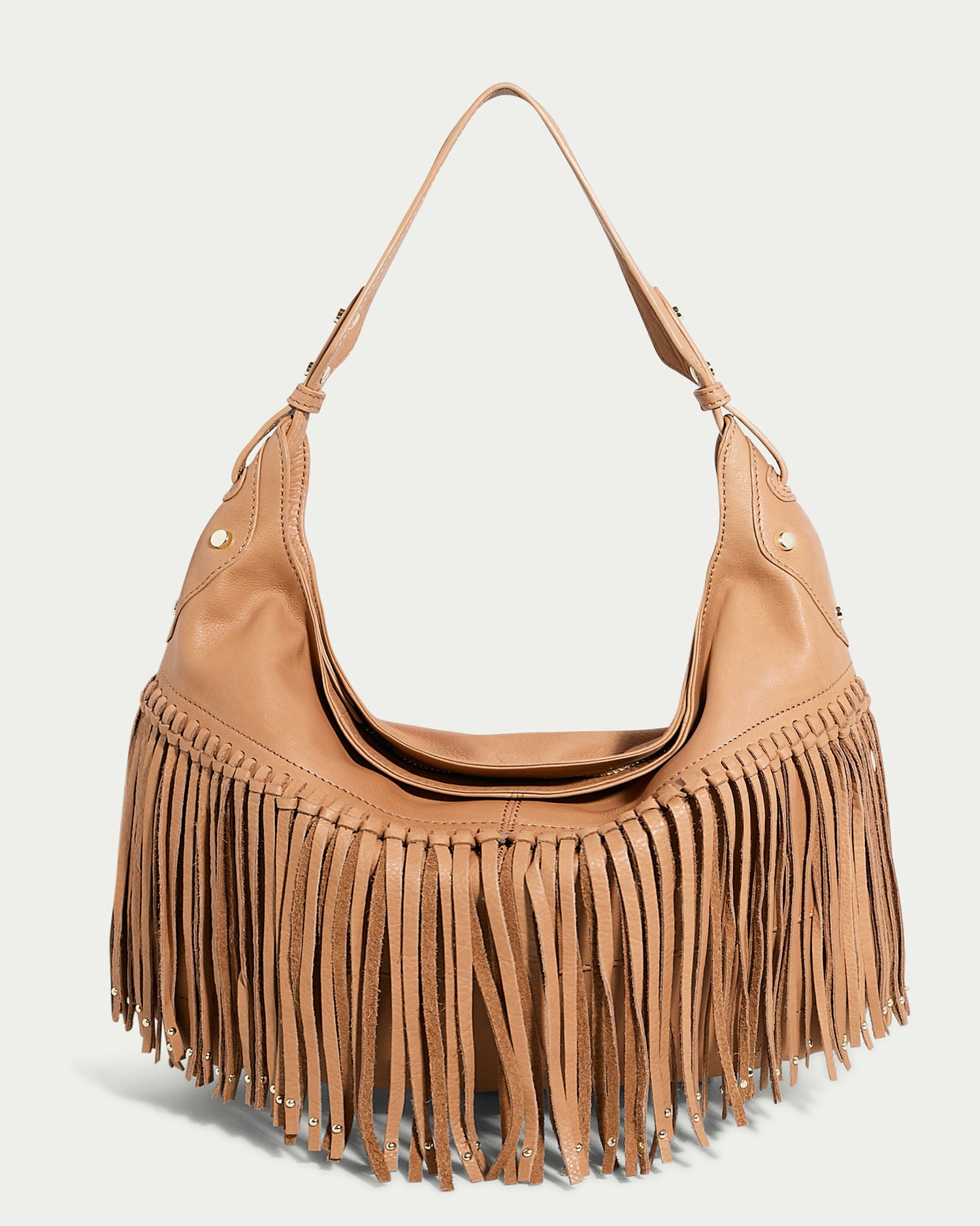 The Lilly Ziptop Hobo from American Leather Co. is crafted from tan genuine leather and features a single shoulder strap along with decorative fringe detailing hanging from the bottom half. Reminiscent of a classic hobo style, this bag includes metal studs on the sides for added embellishment, while its plain, light-colored background highlights its intricate design.