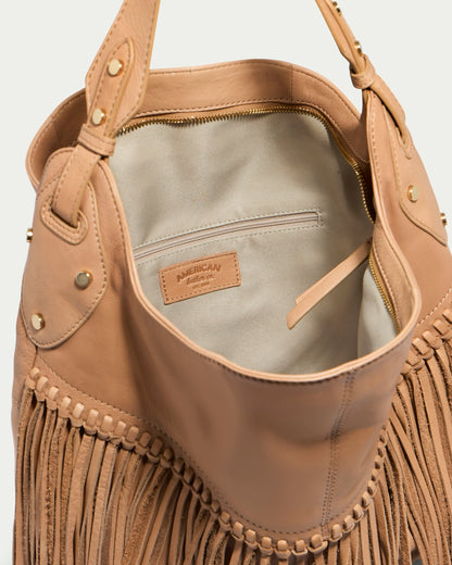 The Lilly Ziptop Hobo, a beige genuine leather handbag with fringe detailing, is displayed open to reveal its light grey interior and a small zippered pocket. The brand label inside reads "American Leather Co." The bag features sturdy handles accented with brass rivets.