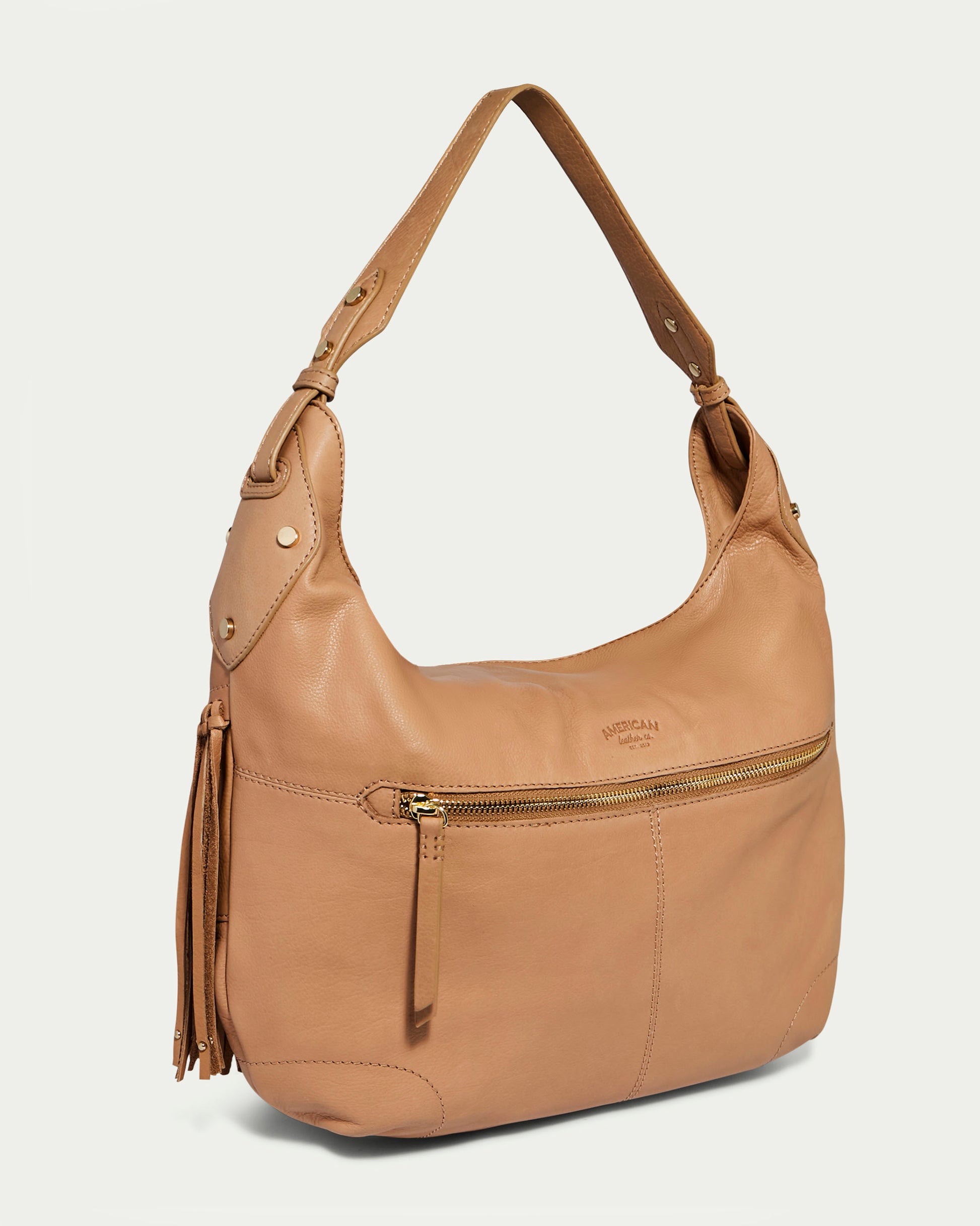 The Lilly Ziptop Hobo by American Leather Co. is a genuine leather tan hobo handbag featuring a single, wide shoulder strap. It boasts a front zippered pocket with a tassel pull, visible stitching, and metal rivet accents, while the main body of the bag has a smooth texture and an embossed subtle brand logo above the zipper.