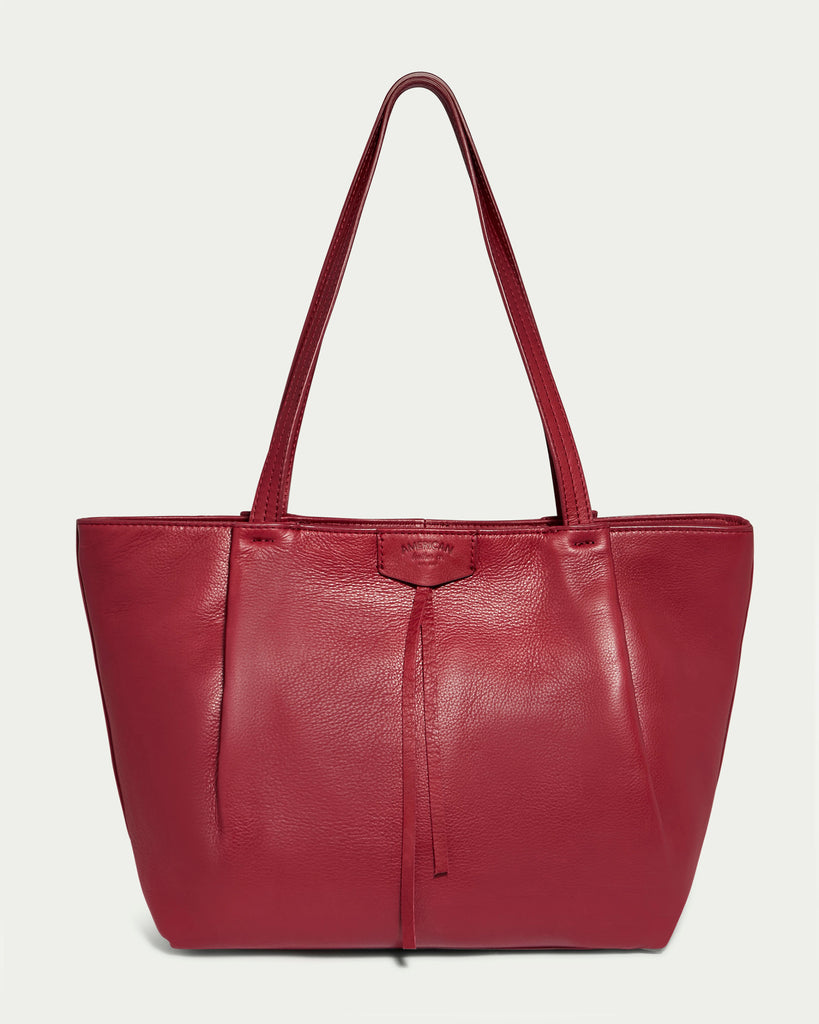 The Lindley Tote by American Leather Co. is a red genuine leather tote bag with dual shoulder straps and a minimalist design. This fashion tote features a slightly textured surface and a small tassel hanging at the center, set against a white background.