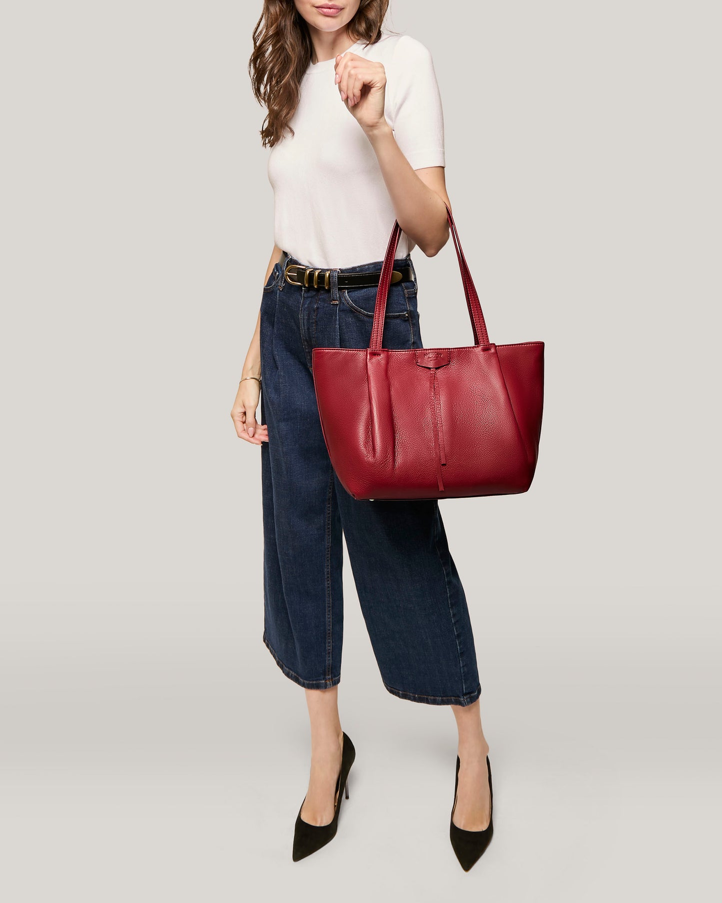 A woman dressed in a white short-sleeve top and dark wide-leg jeans holds the oversized Lindley Tote by American Leather Co. in red leather on her shoulder. She wears black pointed-toe heels and stands against a plain light grey background.