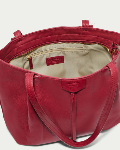 The Lindley Tote is an oversized red leather bag from American Leather Co., featuring dual handles. Its open interior reveals a beige lining and a zippered pocket with a leather pull-tab. The genuine leather craftsmanship is highlighted by the brand name visible on a leather tag both inside and near the top of the bag's exterior.