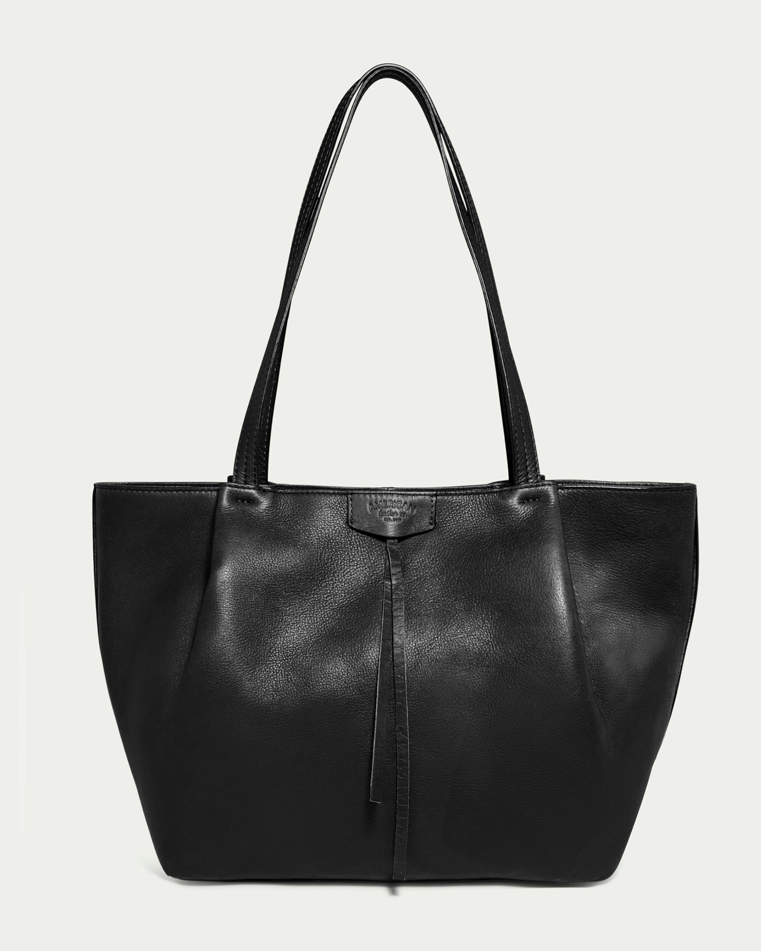 The Lindley Tote by American Leather Co. is a sleek, black oversized bag crafted from genuine leather. It features two long straps with padded sections for comfort, a slightly textured surface, and a minimalistic design. A small central leather tag adorns the top and a single decorative leather strip hangs down the front.