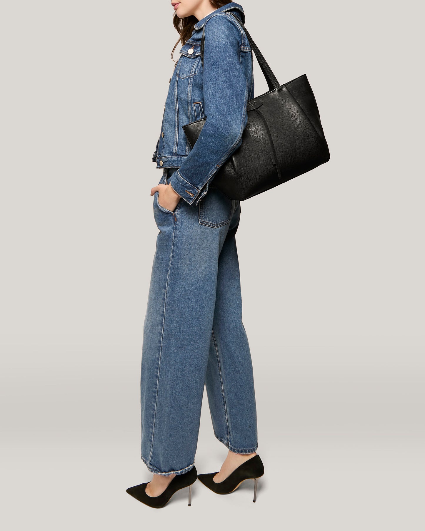 A person is standing against a plain background, wearing a denim jacket and wide-leg jeans. They are carrying the Lindley Tote by American Leather Co., which has padded straps, and are wearing black high-heeled shoes. The outfit has a casual yet chic style. The person's face is not visible in the image.