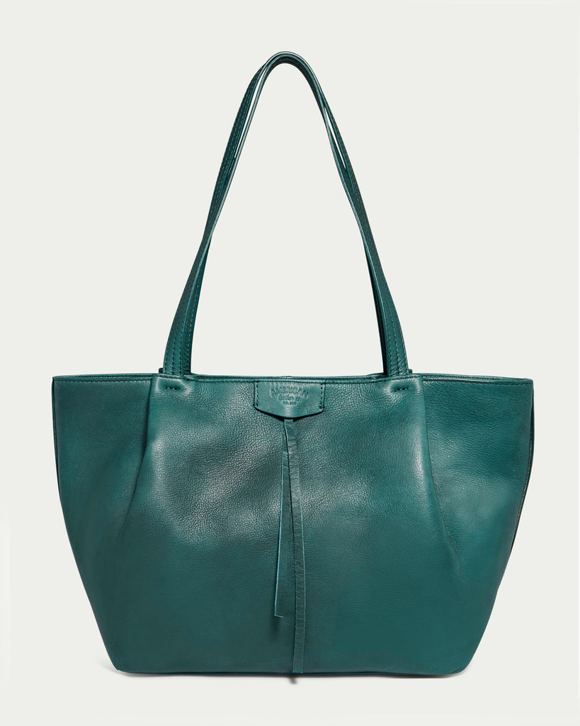 The Lindley Tote by American Leather Co. is a deep green, oversized bag featuring double handles and a minimalist design. The front showcases a small tab detail with a slender strip of matching genuine leather hanging down. This tote has a structured shape and smooth finish.