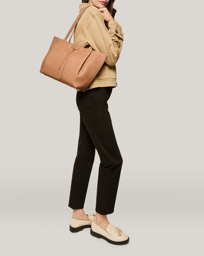 A woman is standing against a plain background, sporting a light brown jacket, black pants, and beige loafers. She has her hands in her jacket pockets and is carrying the American Leather Co. Lindley Tote—an oversized tote with genuine leather and a top zipper closure—on her shoulder.