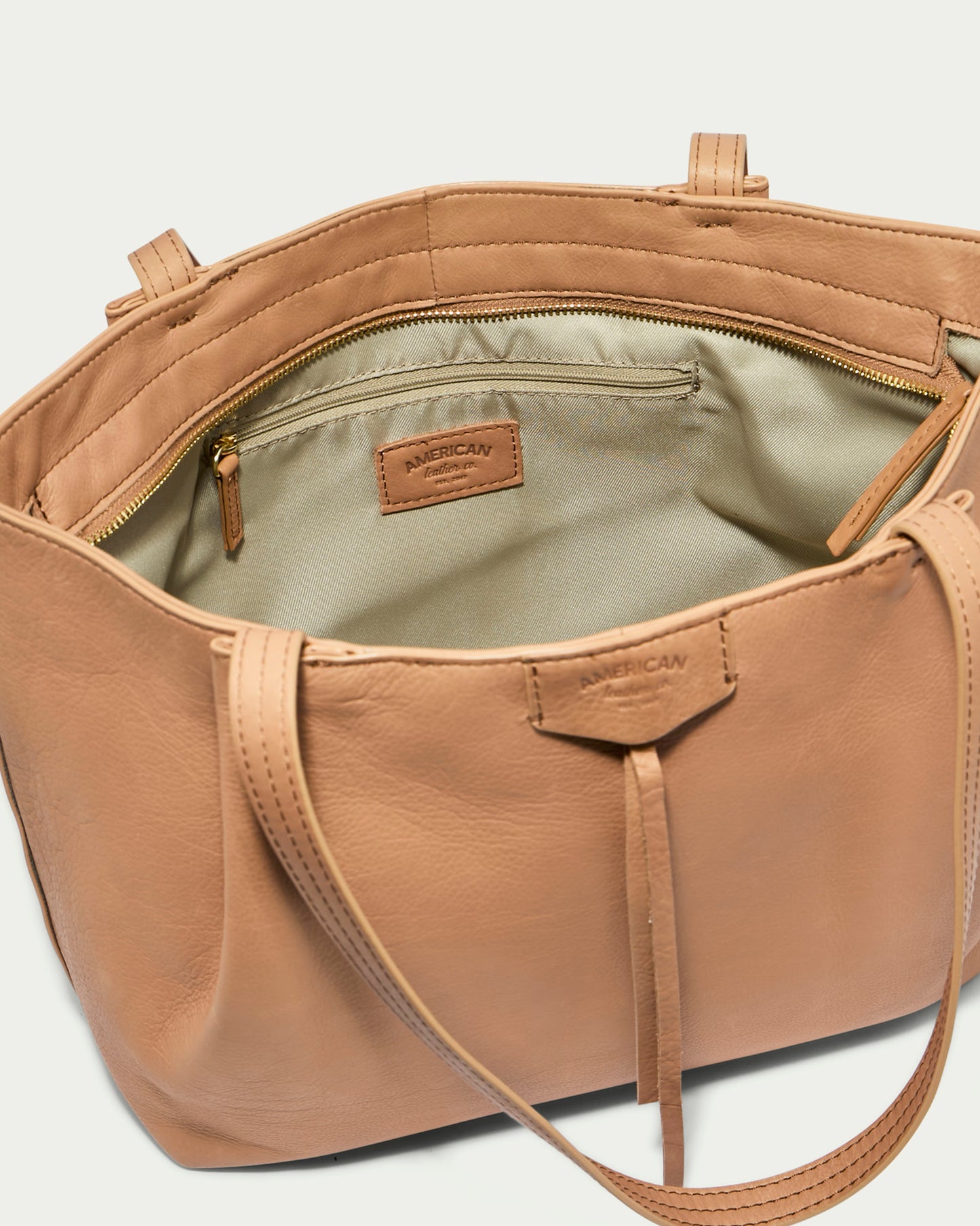 The Lindley Tote by American Leather Co. is a tan genuine leather bag featuring an open top that reveals a beige interior lining. Inside, there's a zippered pocket with an attached leather brand label. This oversized tote comes with dual handles and has a decorative leather tie on the front.