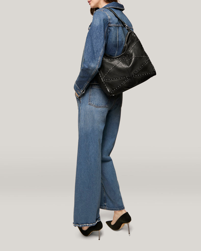 A person stands against a neutral background, wearing a blue denim jacket and wide-legged jeans. They carry the Locke Ziptop Studded Hobo from American Leather Co., and wear black high-heeled shoes. The image shows them from the side, facing slightly away from the camera.