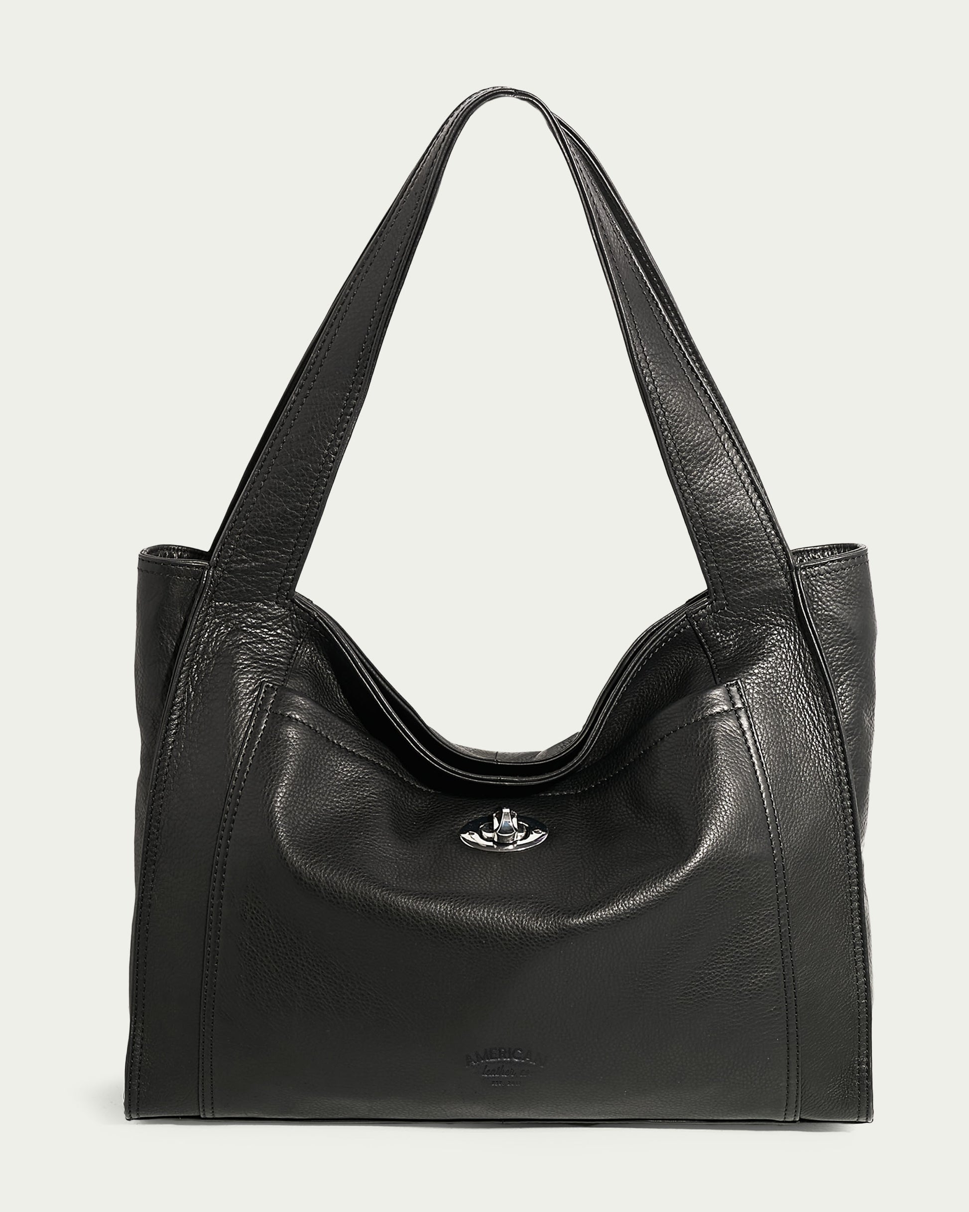 The Plymouth Ziptop Tote by American Leather Co. features a smooth black leather exterior with two wide straps, a silver clasp, and a front pocket. Its structured build is complemented by detailed stitching along the edges, making this genuine leather handbag stand out against the light cream background.