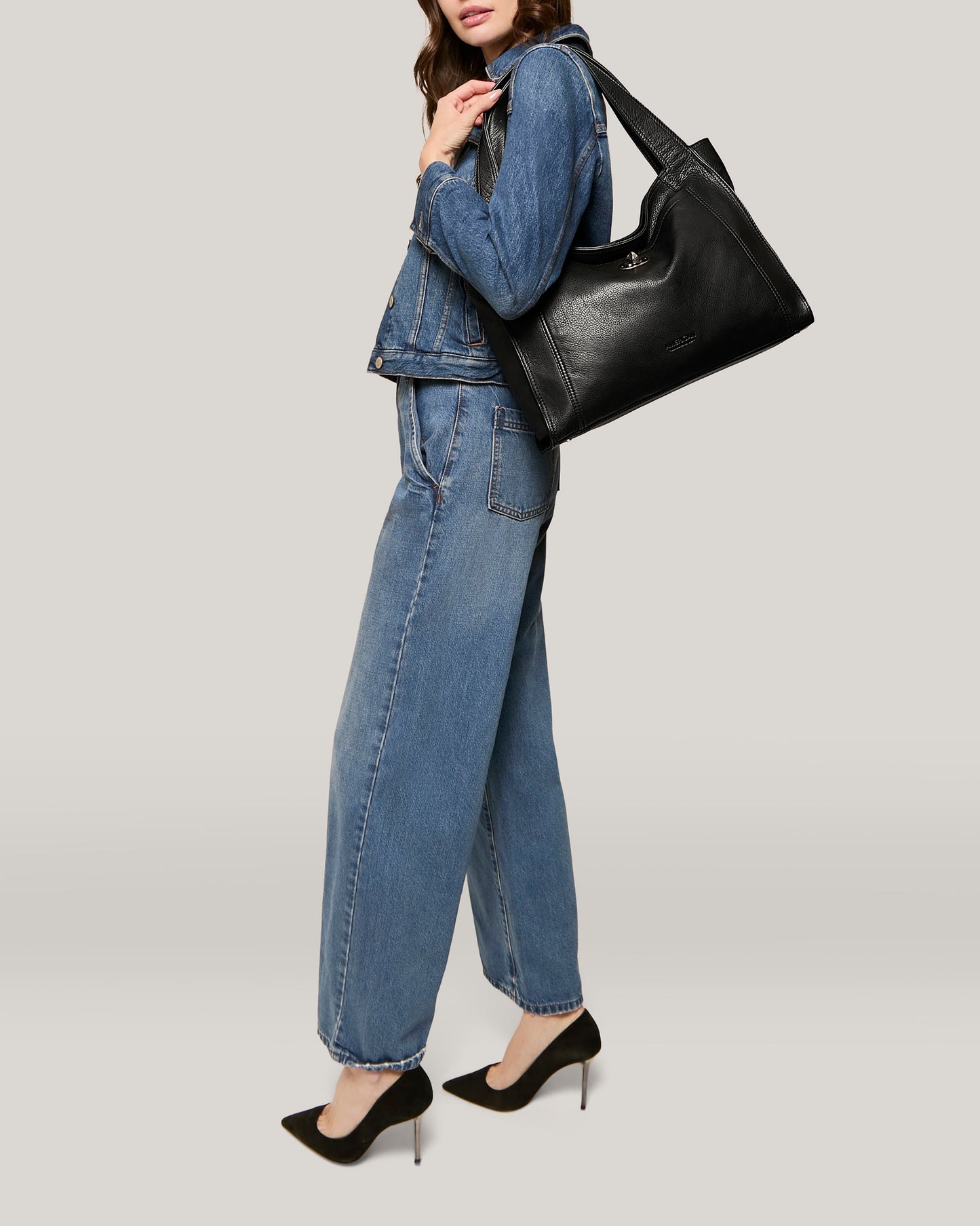 A person is standing against a plain background, dressed in a blue denim jacket and wide-legged jeans. They carry an American Leather Co. Plymouth Ziptop Tote on their shoulder and wear black high-heeled shoes. The person's face is not visible.