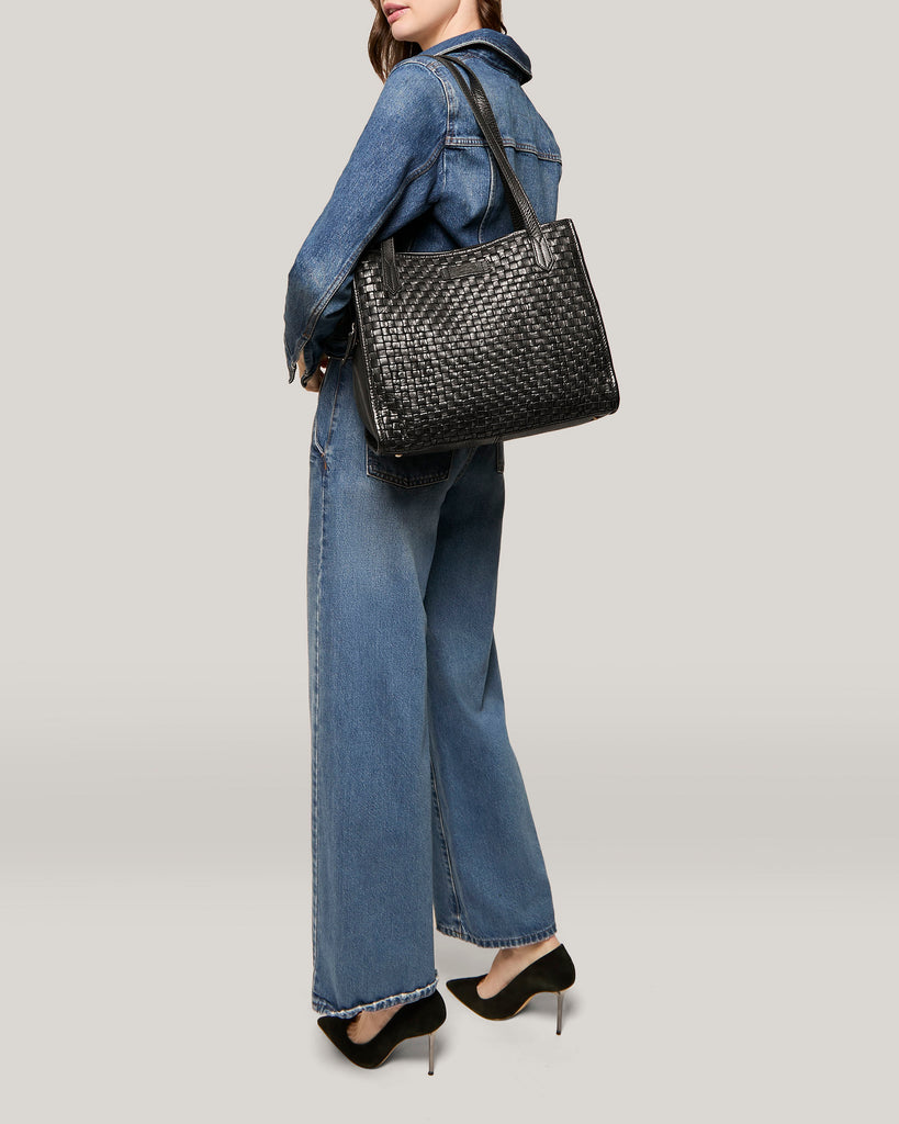 A person wearing a blue denim jacket and wide-leg jeans stands with one hand in their pocket. They carry an American Leather Co. Sandy Woven Triple Entry Satchel on their shoulder, made from genuine leather. The person is wearing black high-heeled shoes. The background is a plain, light gray color.