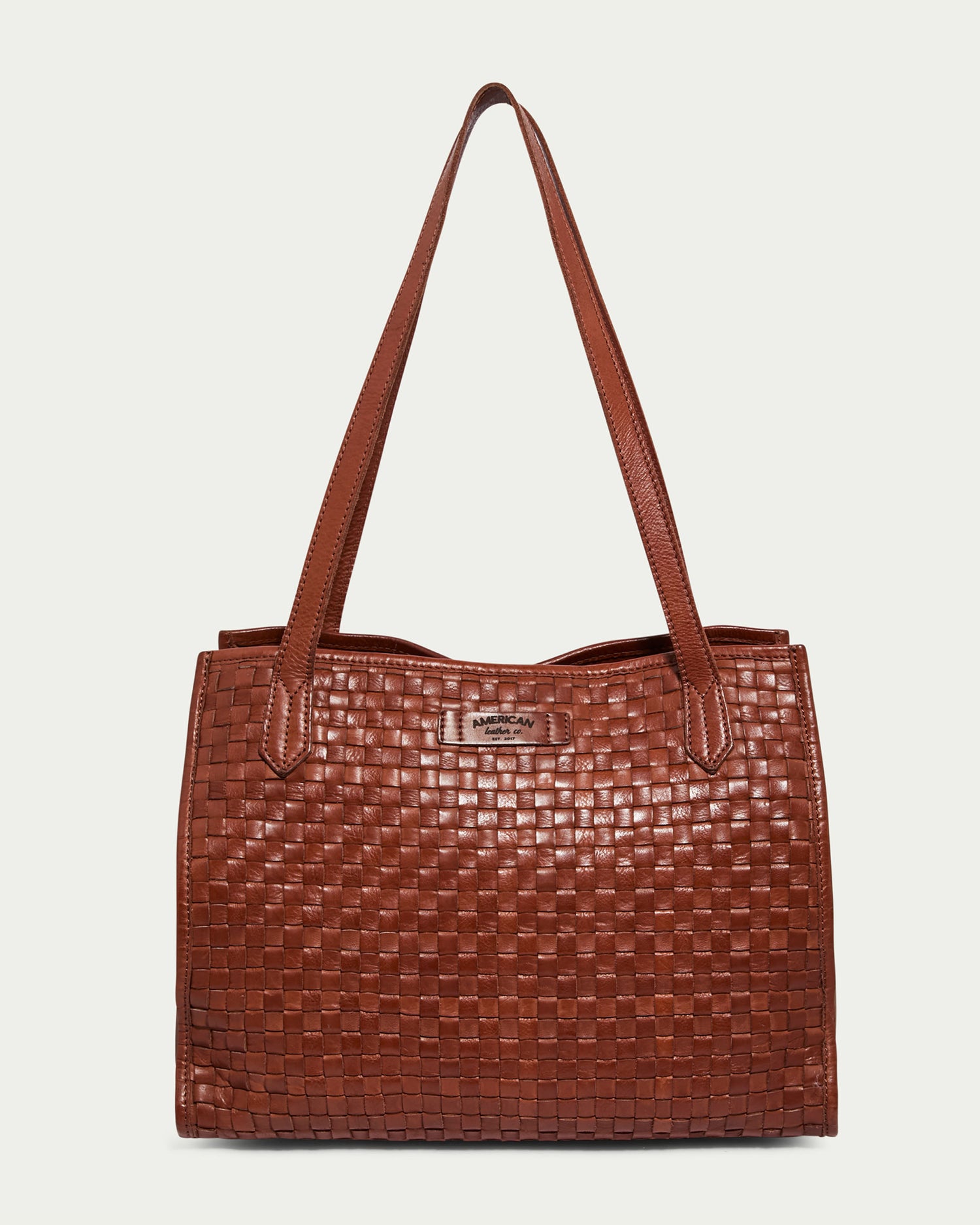 The Sandy Woven Triple Entry Satchel by American Leather Co. in a brandy color features a checkered pattern on its surface and small metallic brand label on the front near the top, along with dual shoulder straps. The background is a plain, light color.