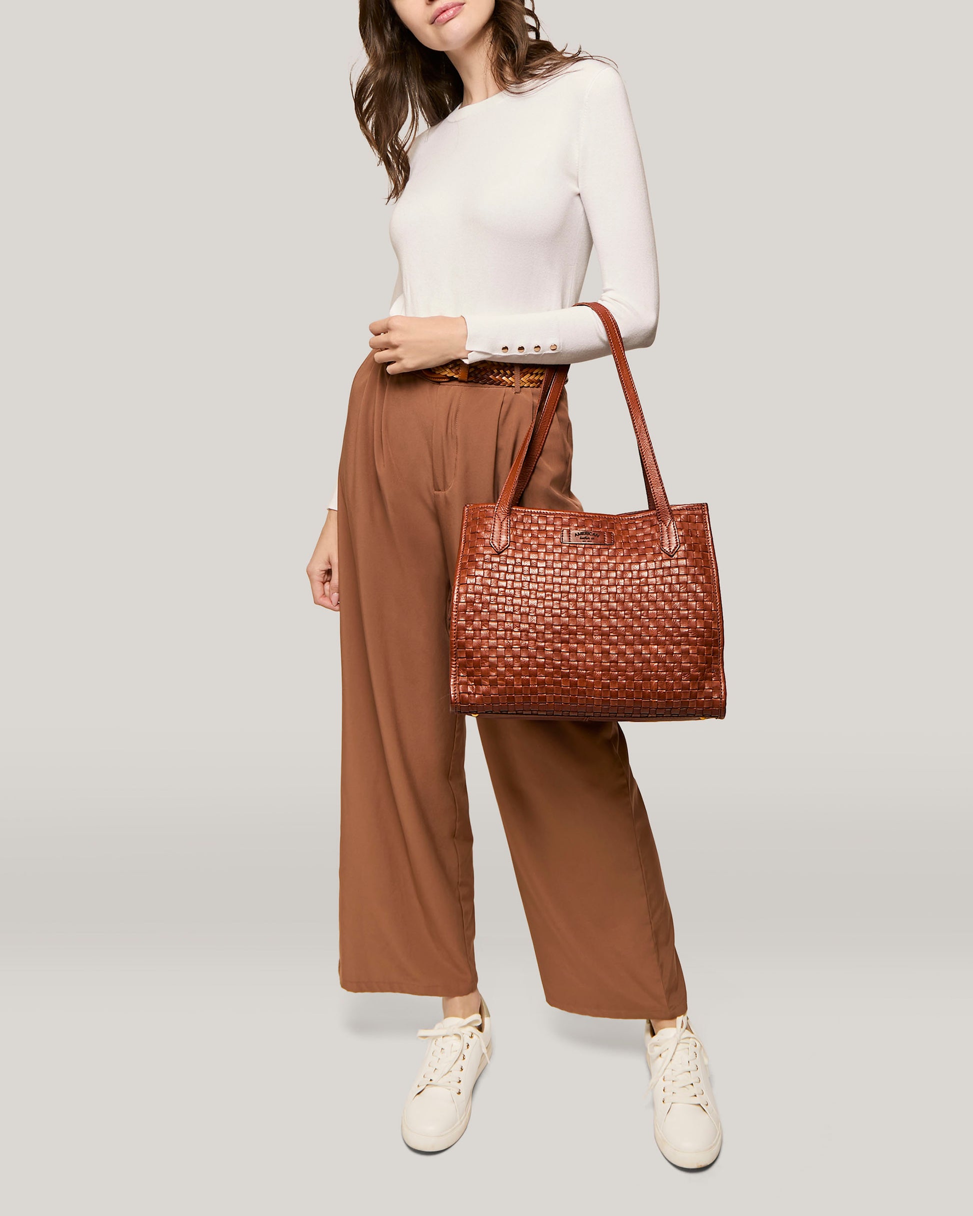 A person wearing a long-sleeve white top, brown high-waisted wide-leg pants, and white sneakers is holding an American Leather Co. Sandy Woven Triple Entry Satchel in their left hand. The background is plain, putting the focus entirely on the outfit.