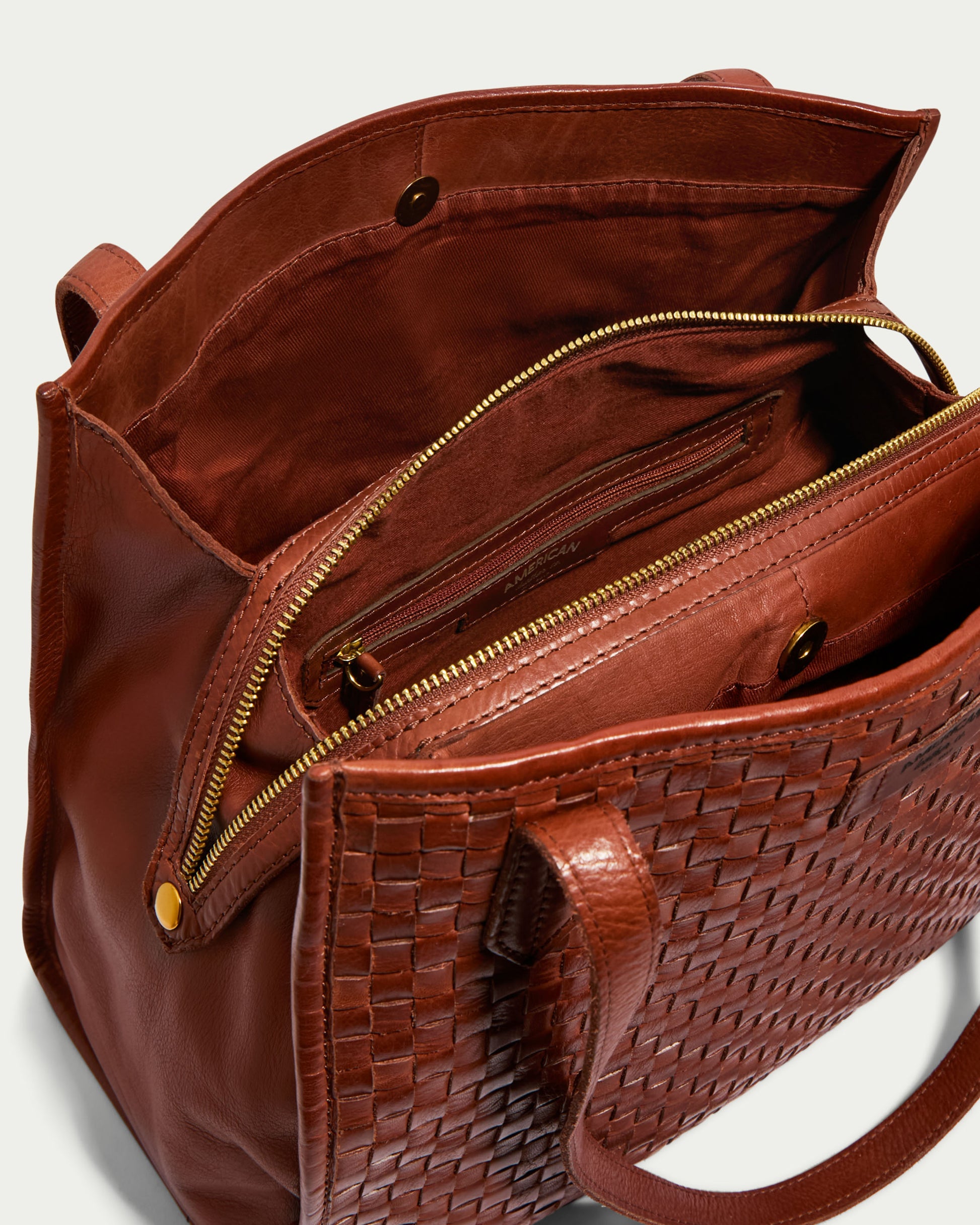 A close-up view of an open brown leather Sandy Woven Triple Entry Satchel showcasing its intricate woven detailing. This American Leather Co. bag features gold-tone zippers and buttons, multiple compartments inside, and double handles. The interior includes a main zippered section, a smaller zippered pocket, and an open pocket with a button closure.