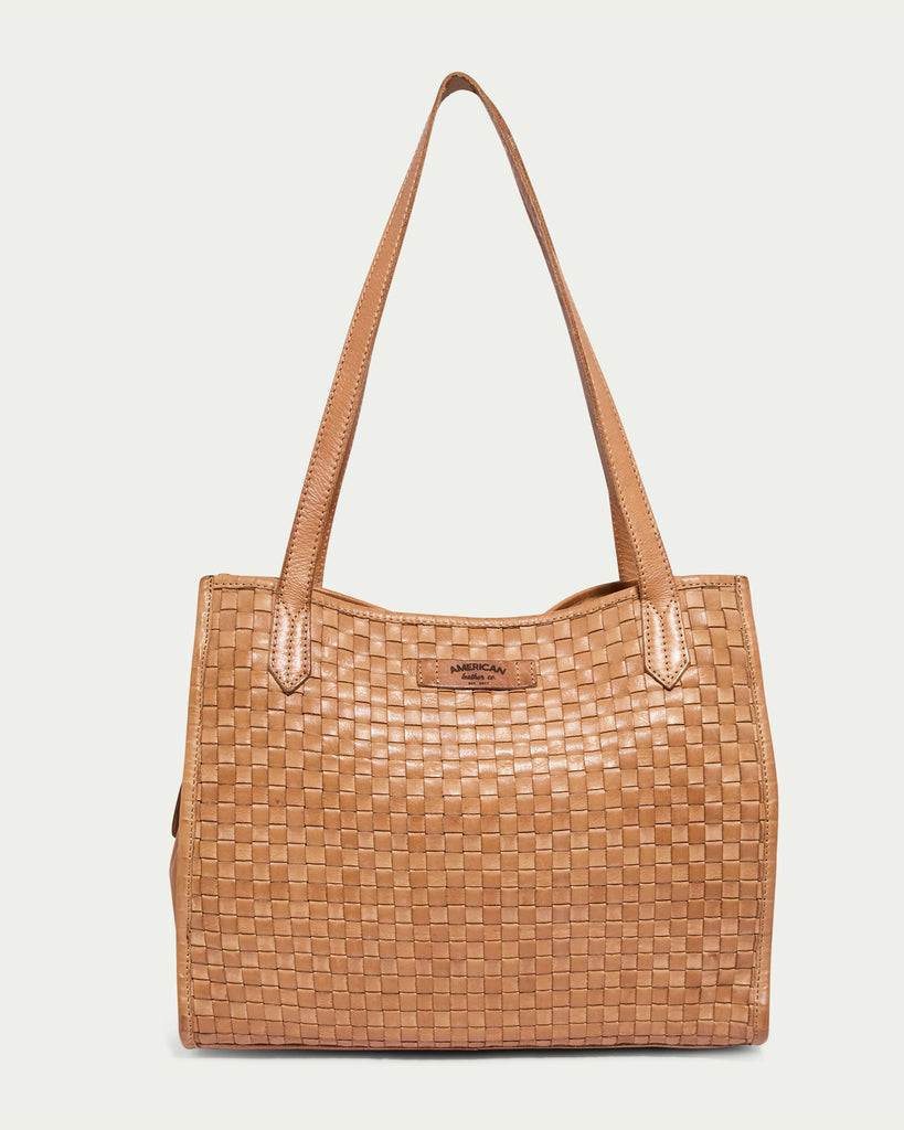 The nutmeg-colored leather handbag, named the Sandy Woven Triple Entry Satchel from American Leather Co., features a woven texture and dual shoulder straps. The rectangular-shaped bag boasts a small tag on the front displaying the brand name and is crafted from genuine leather, set against a plain, light-colored background.