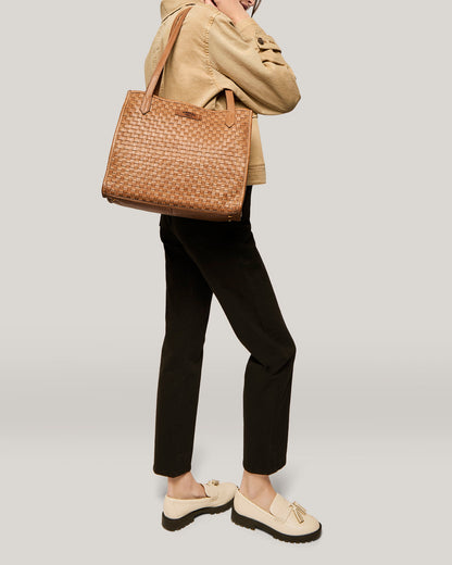 A person is modeling a brown woven tote bag. They are dressed in a beige jacket, black pants, and white loafers with tassels. The background is plain and light-colored. The focus is on the American Leather Co.'s Sandy Woven Triple Entry Satchel, slung over the person's shoulder.