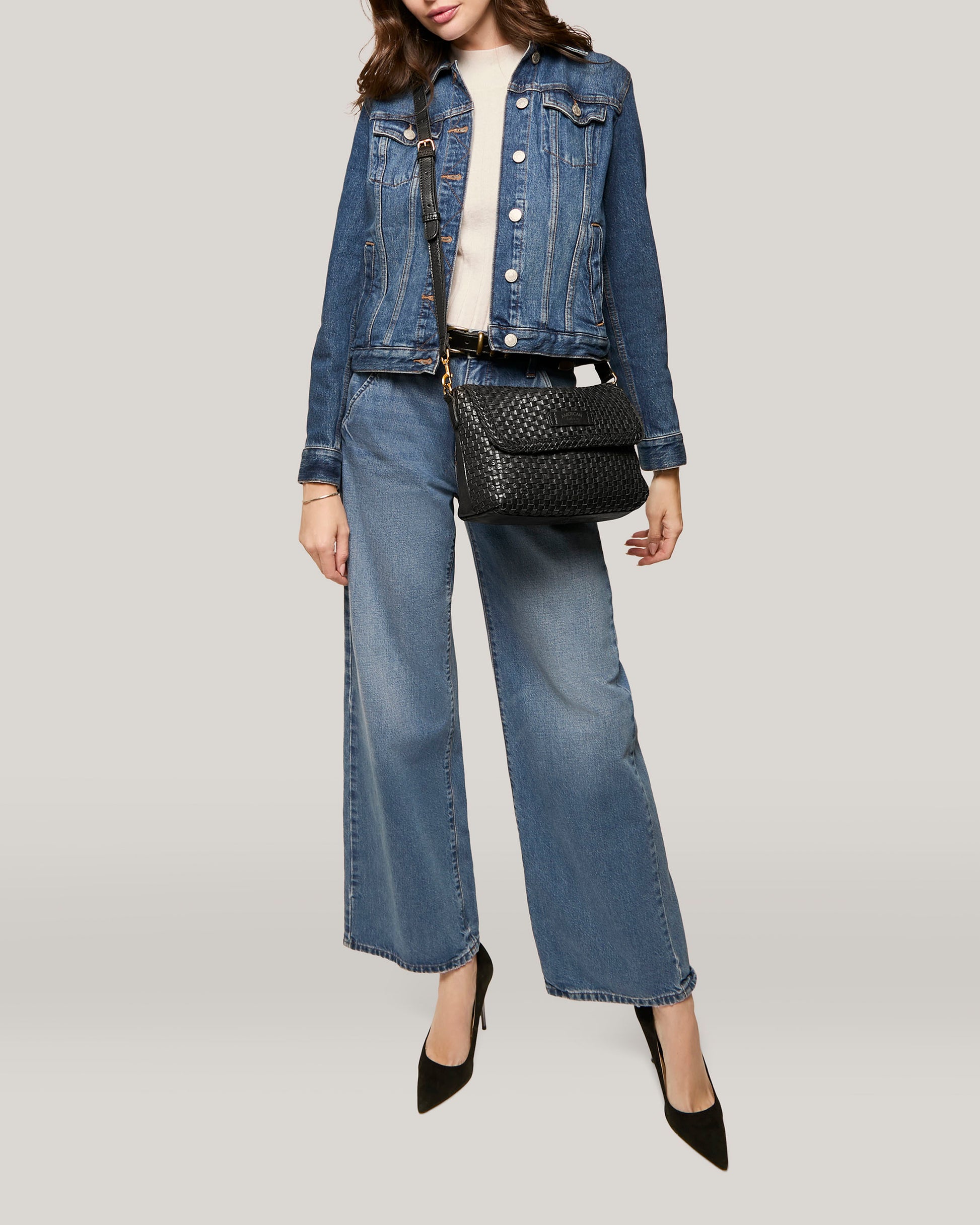 A person is shown from the neck down wearing a blue denim jacket, wide-leg denim jeans, and a light-colored top. They carry the American Leather Co. Summers Woven Crossbody handbag in black and wear black pointed-toe high heels. The background is a plain light gray.