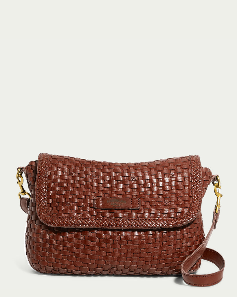 The Summers Woven Crossbody by American Leather Co. is a brown, woven leather bag featuring a flap closure and an adjustable strap with a gold-tone buckle. Its rectangular shape and textured appearance lend it a stylish and sophisticated look against the plain, light-colored background.