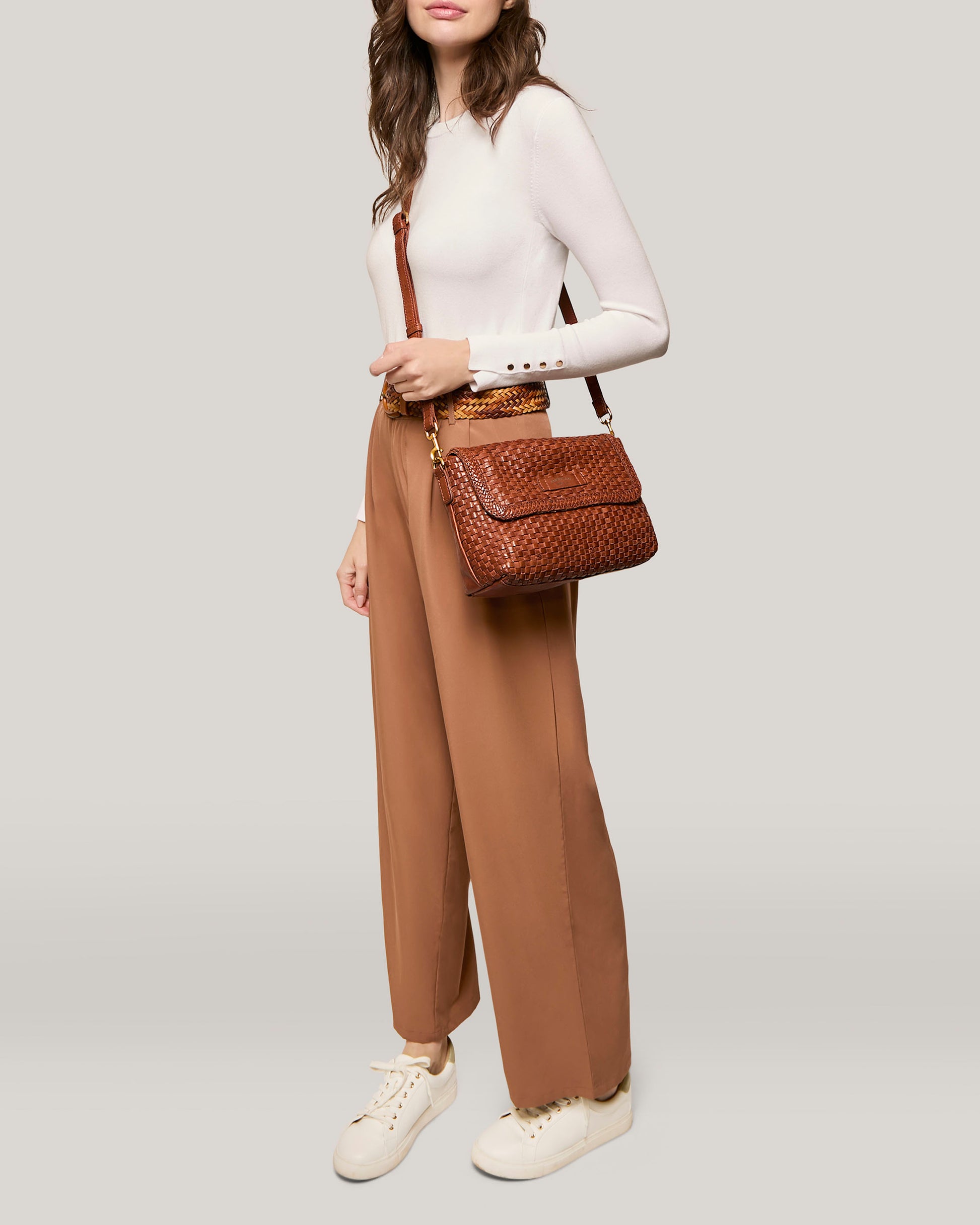 A woman with long hair wears a white long-sleeve top and high-waisted, wide-legged brown trousers. She carries the Summers Woven Crossbody from American Leather Co., a textured genuine leather bag with an adjustable strap, and wears white sneakers. The background is a solid light gray.