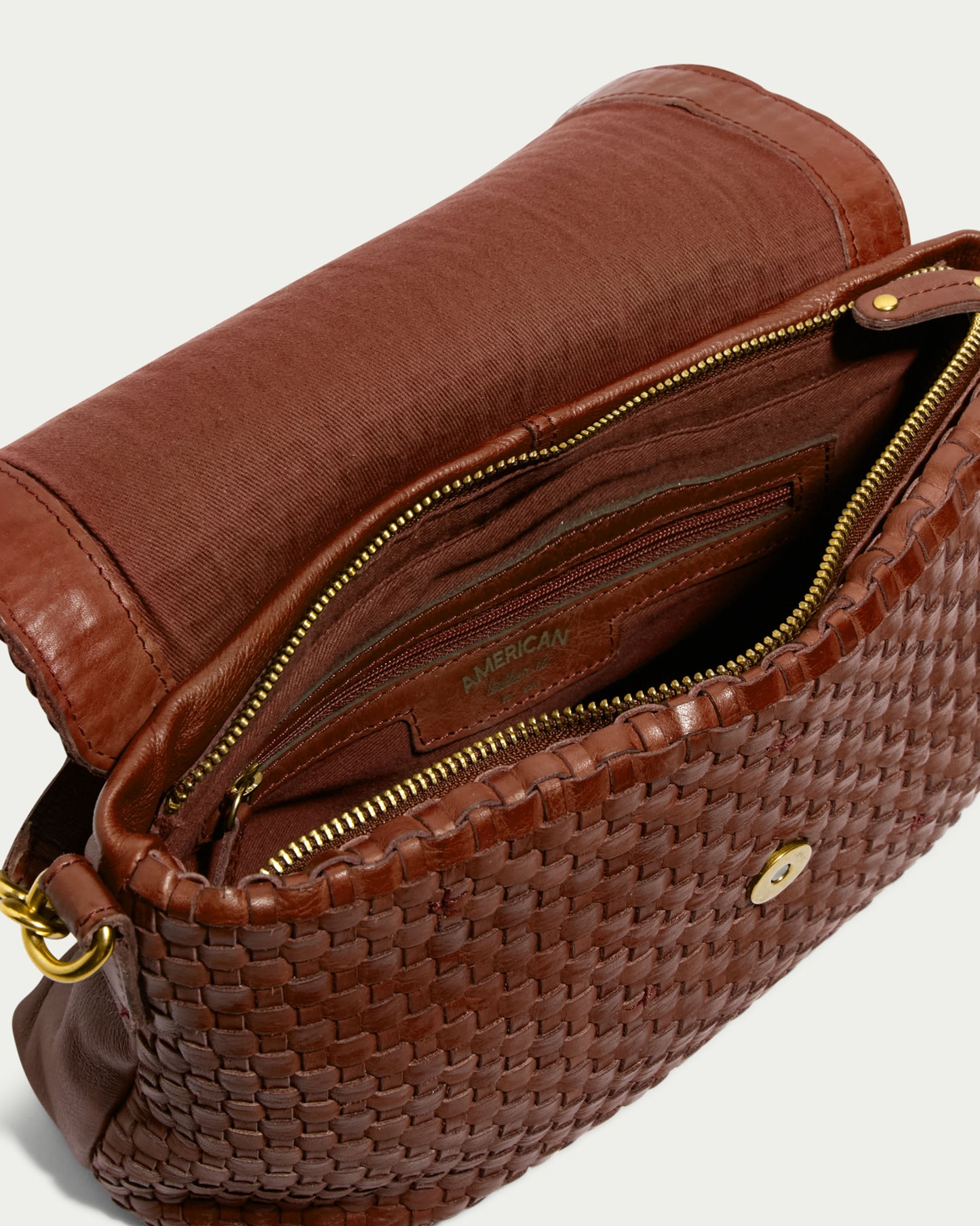 The Summers Woven Crossbody by American Leather Co. is a brown leather crossbody bag with its flap open, revealing a lined interior. Inside, the bag features a secured zipper pocket, open compartments, and an engraved leather label. It has gold-tone hardware, an adjustable strap, and a magnetic snap closure on the flap.