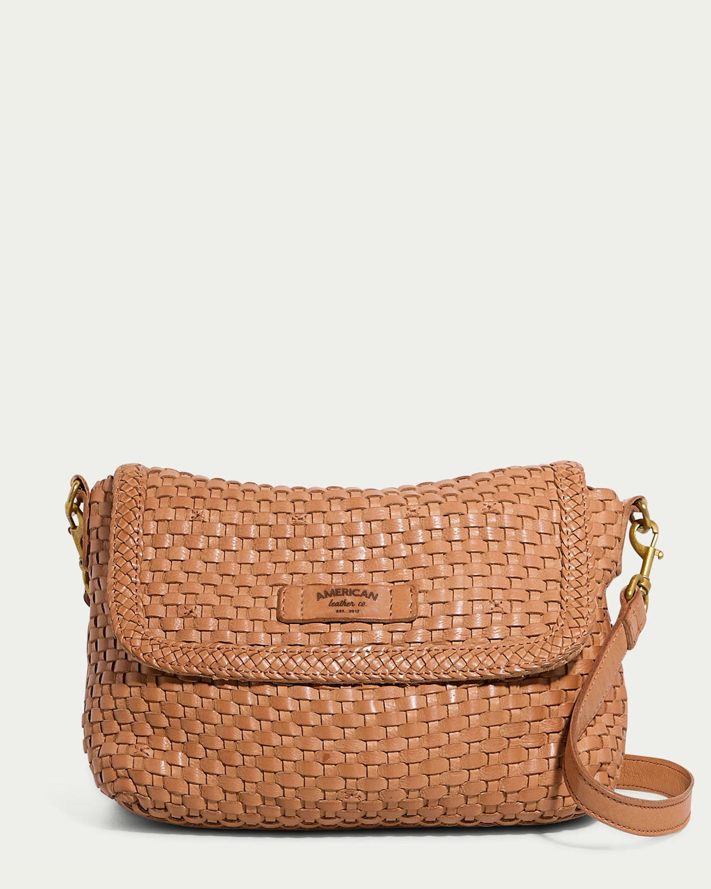 The Summers Woven Crossbody by American Leather Co. is a medium-sized, tan-colored handbag that boasts a woven design and features a flap closure alongside a matching adjustable shoulder strap attached with gold-tone hardware. Crafted from Genuine Leather, the bag displays a small leather patch on the front with text proudly marking it as part of the Heritage collection by American Leather Co.