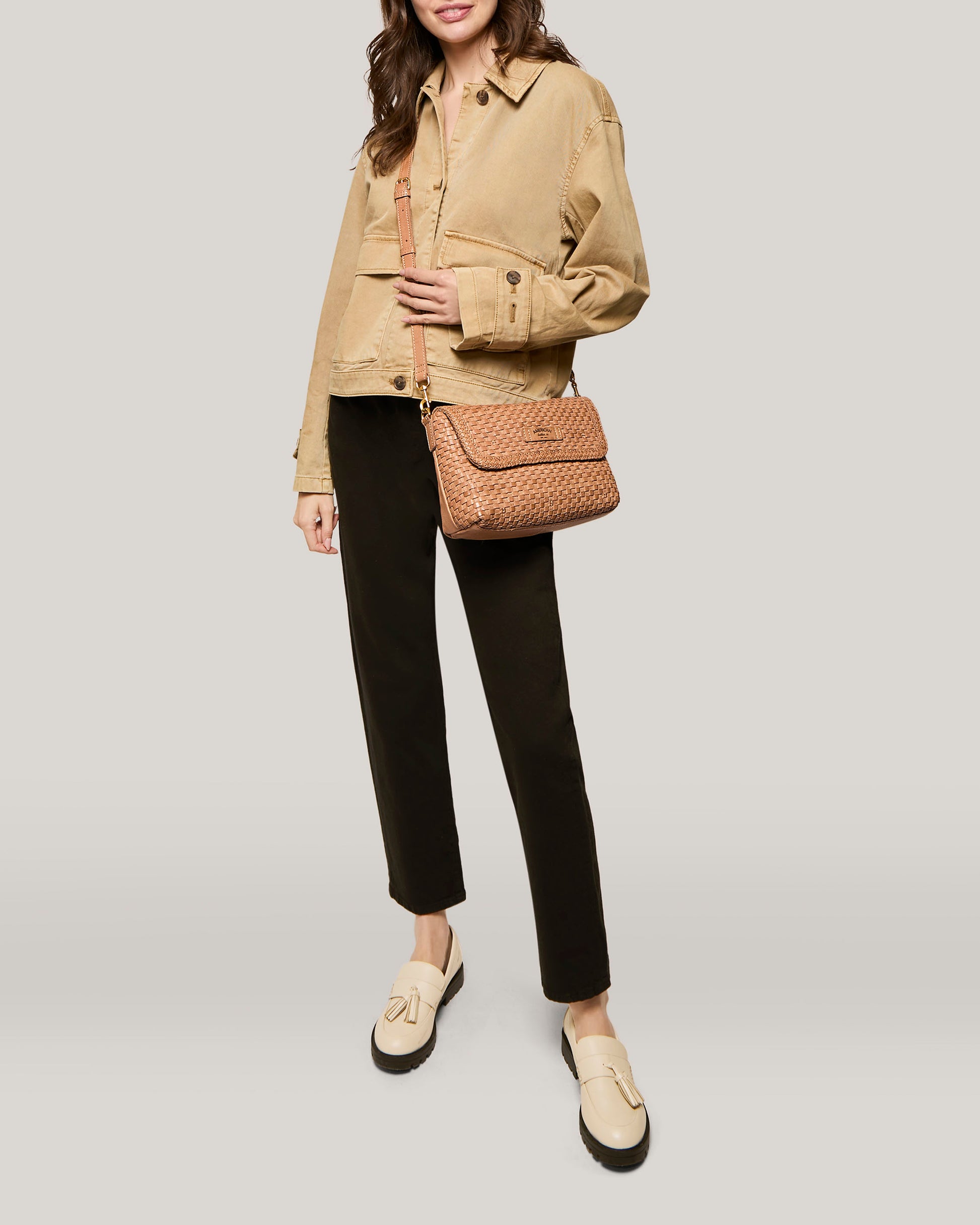 A person wearing a beige jacket and black pants stands against a neutral background. They carry a Summers Woven Crossbody by American Leather Co. over their shoulder and are wearing light-colored loafers.