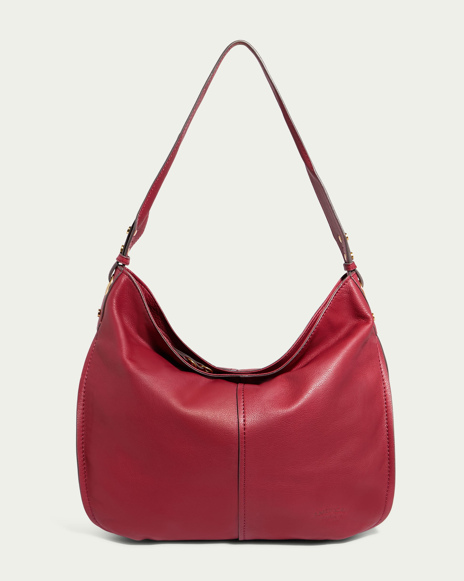 The Summit Large Top Zip Hobo by American Leather Co. is a red genuine leather oversized hobo handbag featuring a single shoulder strap and subtle stitching. It has a rounded, slouchy shape with a minimalist design, includes an adjustable shoulder strap with metallic hardware, and features a zippered main compartment along with a small brand logo embossed on the front.