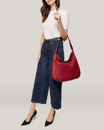 A person with wavy, dark hair wears a white short-sleeve top, high-waisted dark blue wide-leg jeans, and black high-heeled shoes. They carry an American Leather Co. Summit Large Top Zip Hobo handbag over their shoulder that features a spacious zippered main compartment, while their head is not visible in the image.