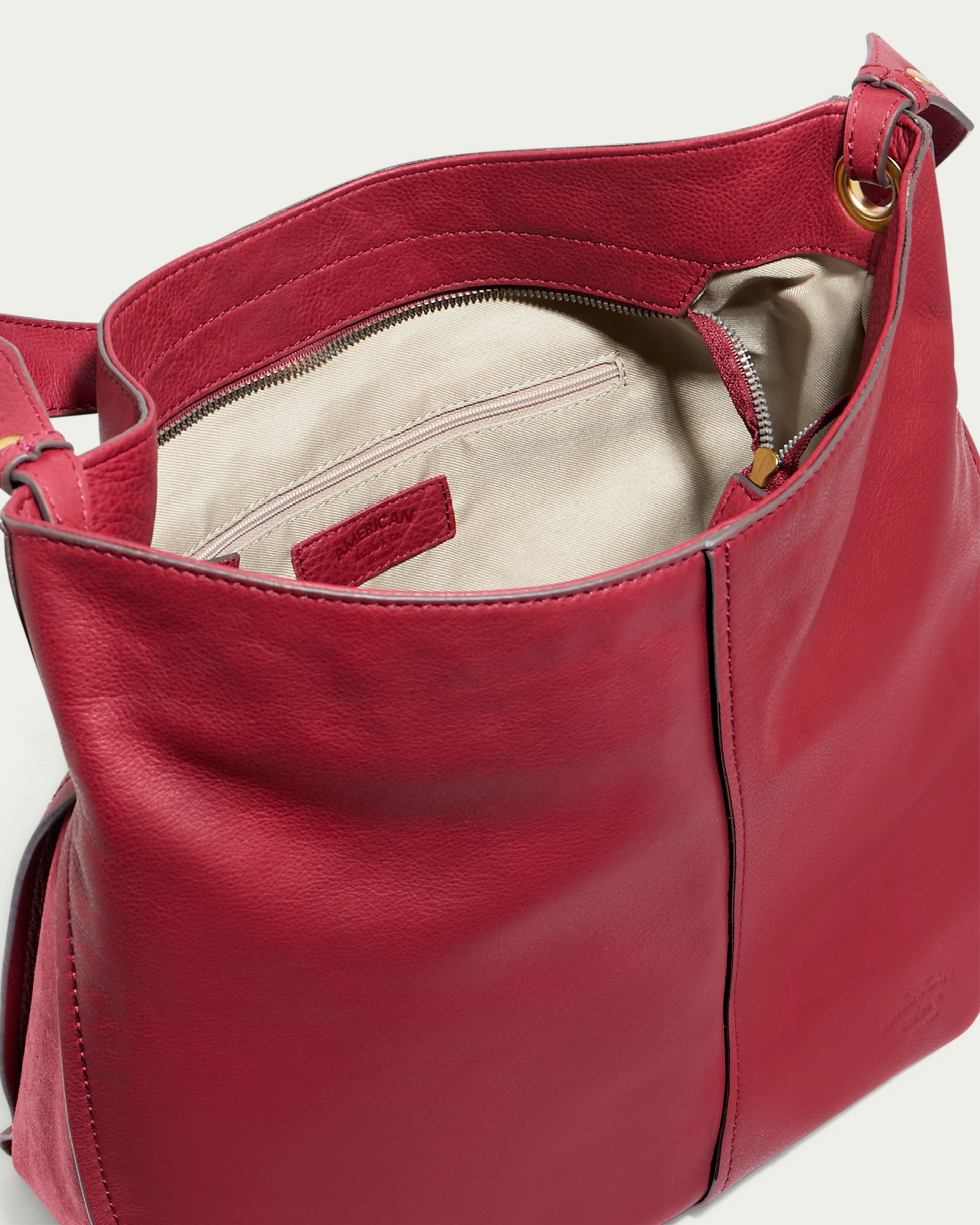 A close-up view of an open Summit Large Top Zip Hobo by American Leather Co., made from genuine red leather. The bag is lined with beige fabric and includes an interior zippered pocket. A gold ring attaches the strap to the bag. Designed with two front panels and a visible seam, it features a zippered main compartment for added security.