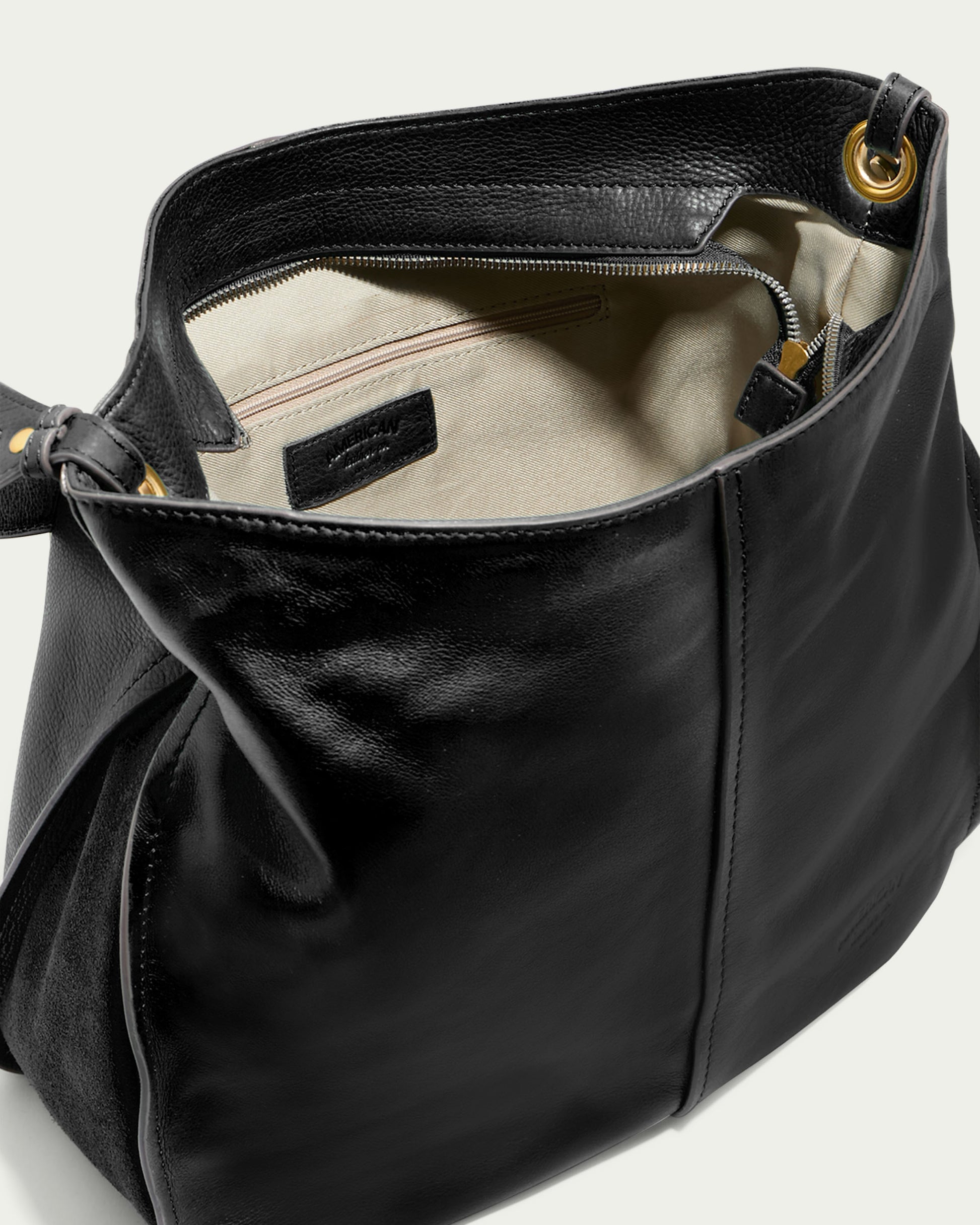 The Summit Large Top Zip Hobo by American Leather Co. is a black genuine leather handbag with a spacious interior featuring a beige lining. It includes an inner zippered pocket and a smaller open pocket, with gold grommet details on the top edges. The bag's leather texture appears smooth and polished, resembling an oversized hobo bag.