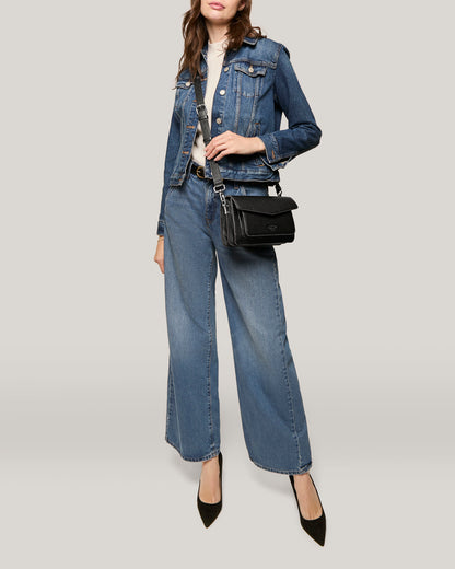 A woman stands against a light grey background, wearing a denim jacket paired with wide-leg denim pants and a white turtleneck. She has an adjustable strap American Leather Co. Troy Acordian Crossbody purse and pointed black heels. Her hair is styled in loose waves.