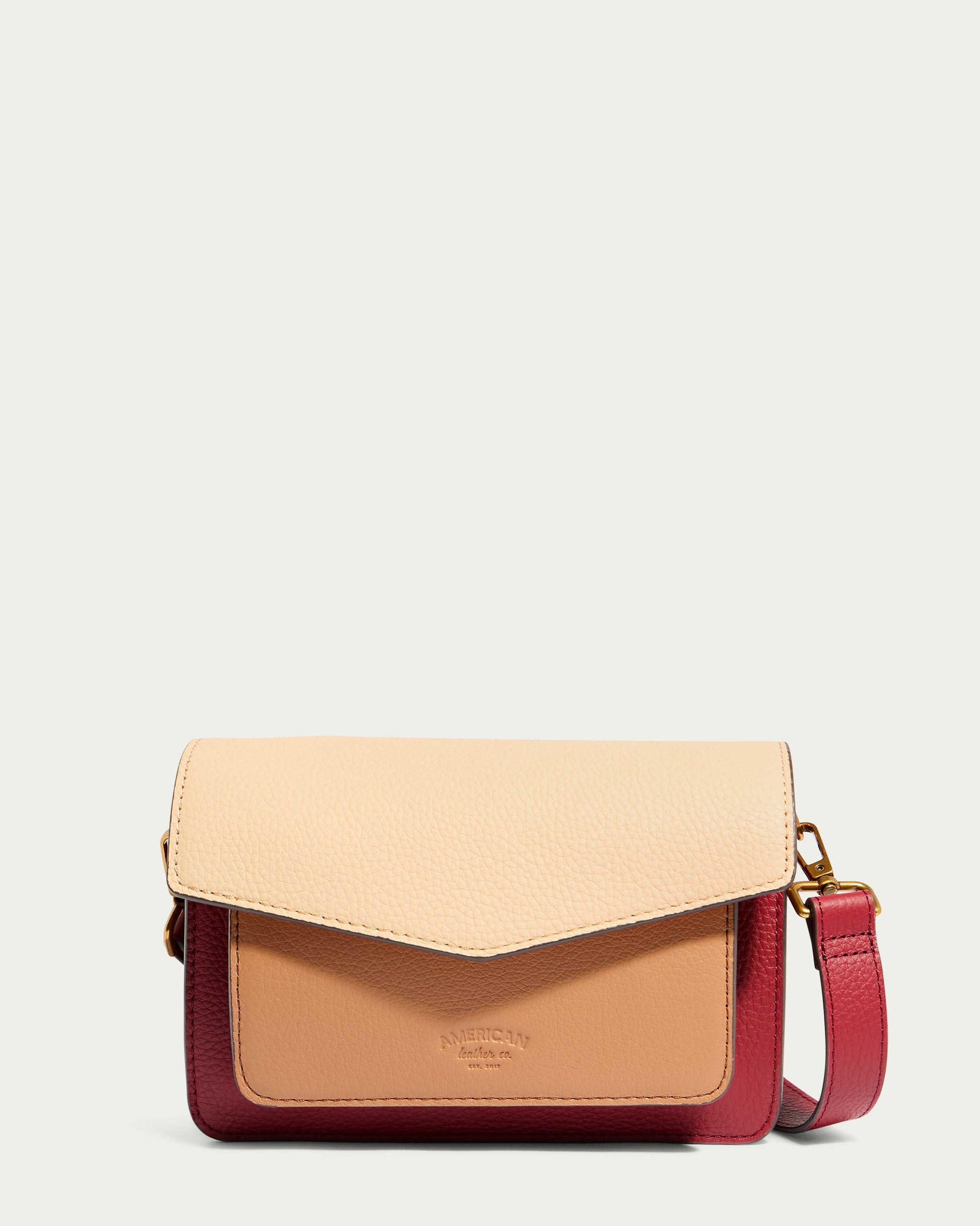 The Troy Acordian Crossbody by American Leather Co. is a fashionable crossbody bag crafted from weatherproof leather, showcasing an elegant color-block design with beige, tan, and red sections. It includes a flap closure, an adjustable red strap, and has the brand name embossed on the front pocket against a light, neutral background.