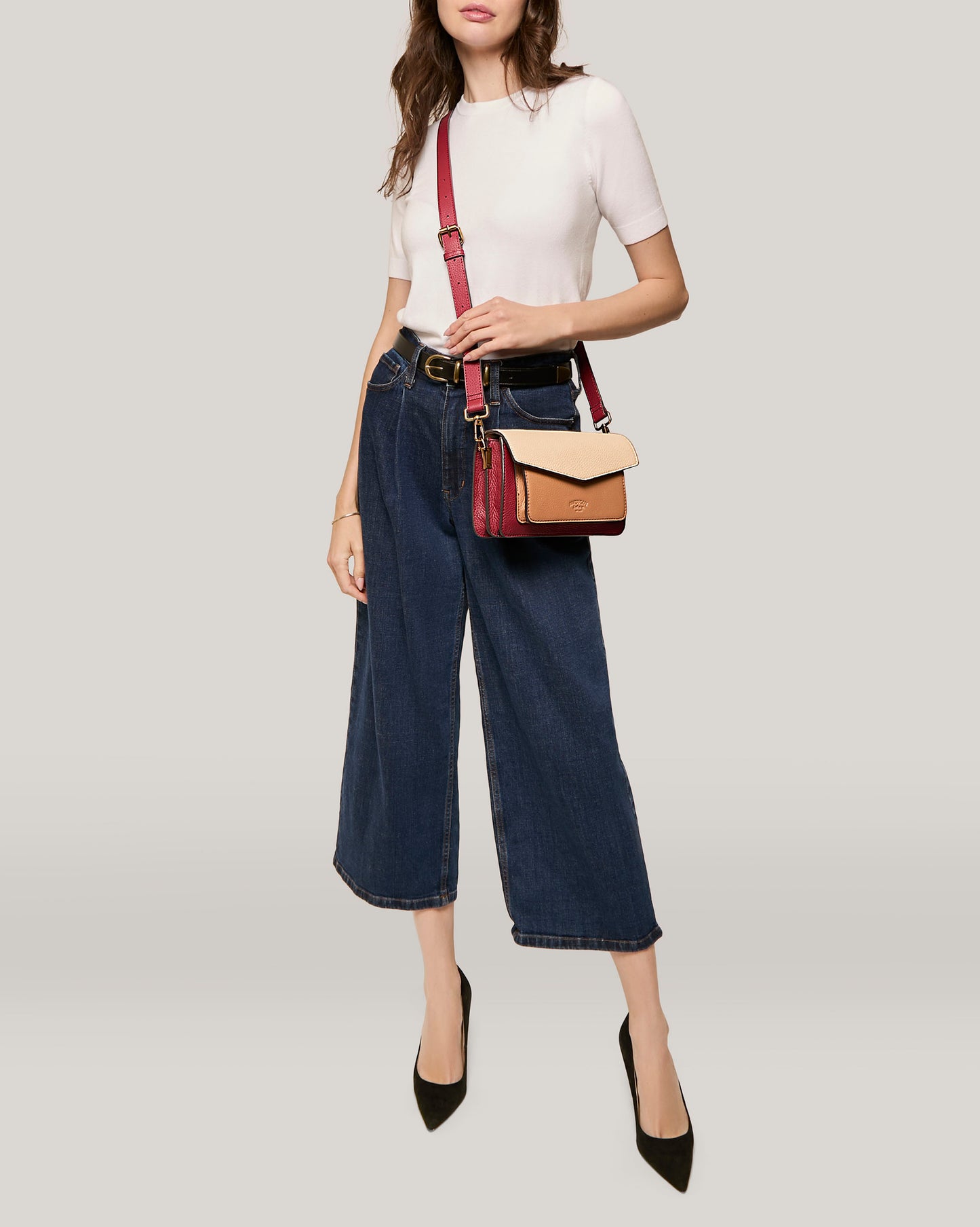 A woman wearing a white short-sleeve top, dark wide-leg jeans, and black heels poses with the Troy Acordian Crossbody by American Leather Co. The background is a light, solid color.