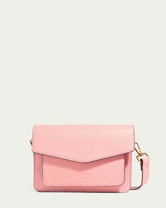 The Troy Acordian Crossbody by American Leather Co. is a stylish pink bag crafted from genuine leather, featuring a fold-over flap and a gold-tone buckle on the side. It boasts an embossed logo on the front and an adjustable strap, combining elegance with versatility.