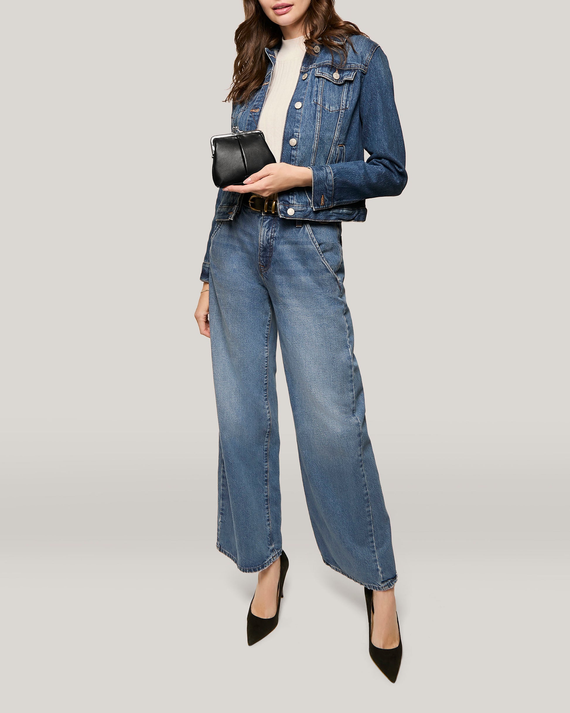 A person dressed in a denim jacket and wide-leg denim jeans holds a small Wolfe Frame Kisslock With CC Slots wallet from the American Leather Co. Cleveland Collection. They are also wearing black pointed-toe heels and a light-colored top, set against a plain, light beige background.