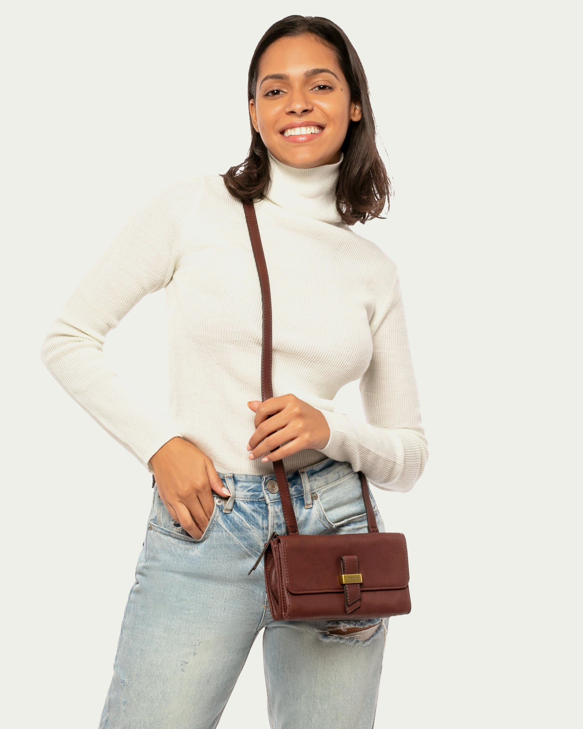 A person in a white turtleneck and light blue jeans smiles while holding an Essex Wallet Crossbody from American Leather Co. The small brown bag, made of genuine leather, features a buckle clasp and adjustable crossbody strap against a plain, light backdrop.