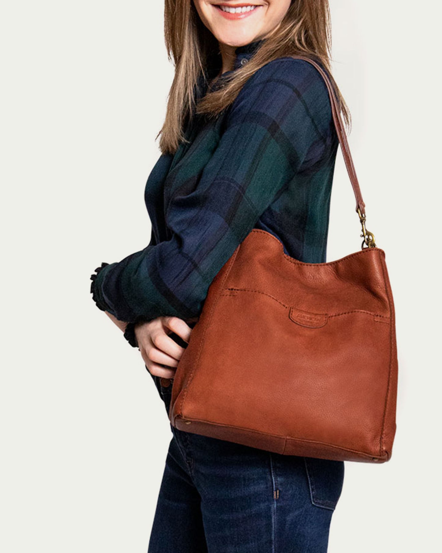 A woman in a plaid shirt and jeans smiles, carrying the American Leather Co. Austin Triple Entry Hobo on her shoulder. This genuine leather bag adds charm against a plain, light-colored background.