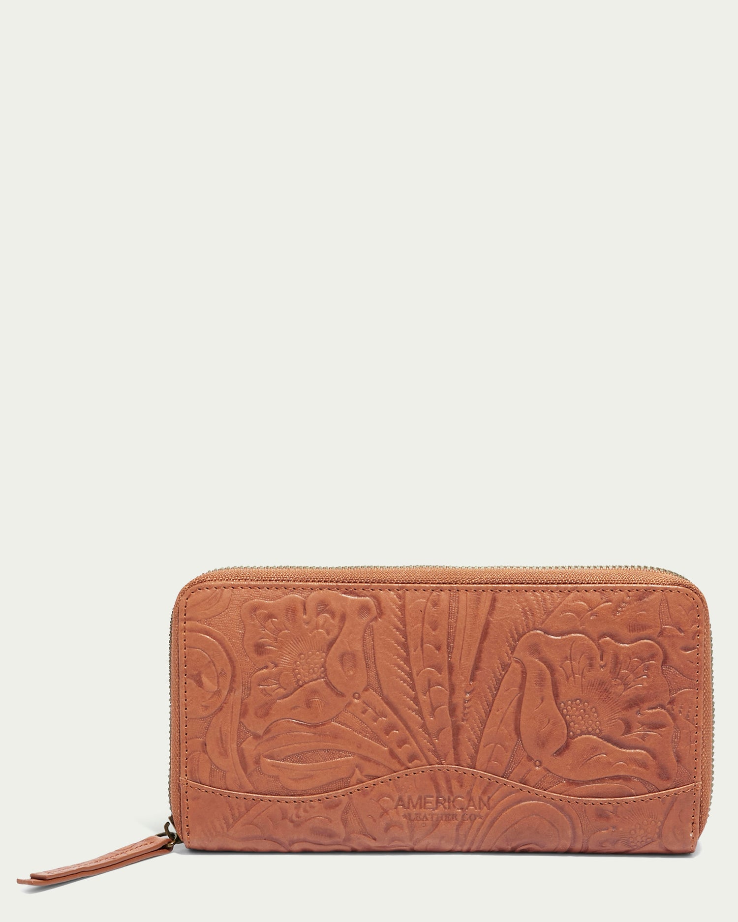 Centered on a plain beige background is the Rye Large Zip Around Wallet from American Leather Co., made from genuine American leather in brown. It features floral engravings and a zipper closure, with detailed embossing of flowers and leaves, complemented by artisanal stitching for an elegant finishing touch.