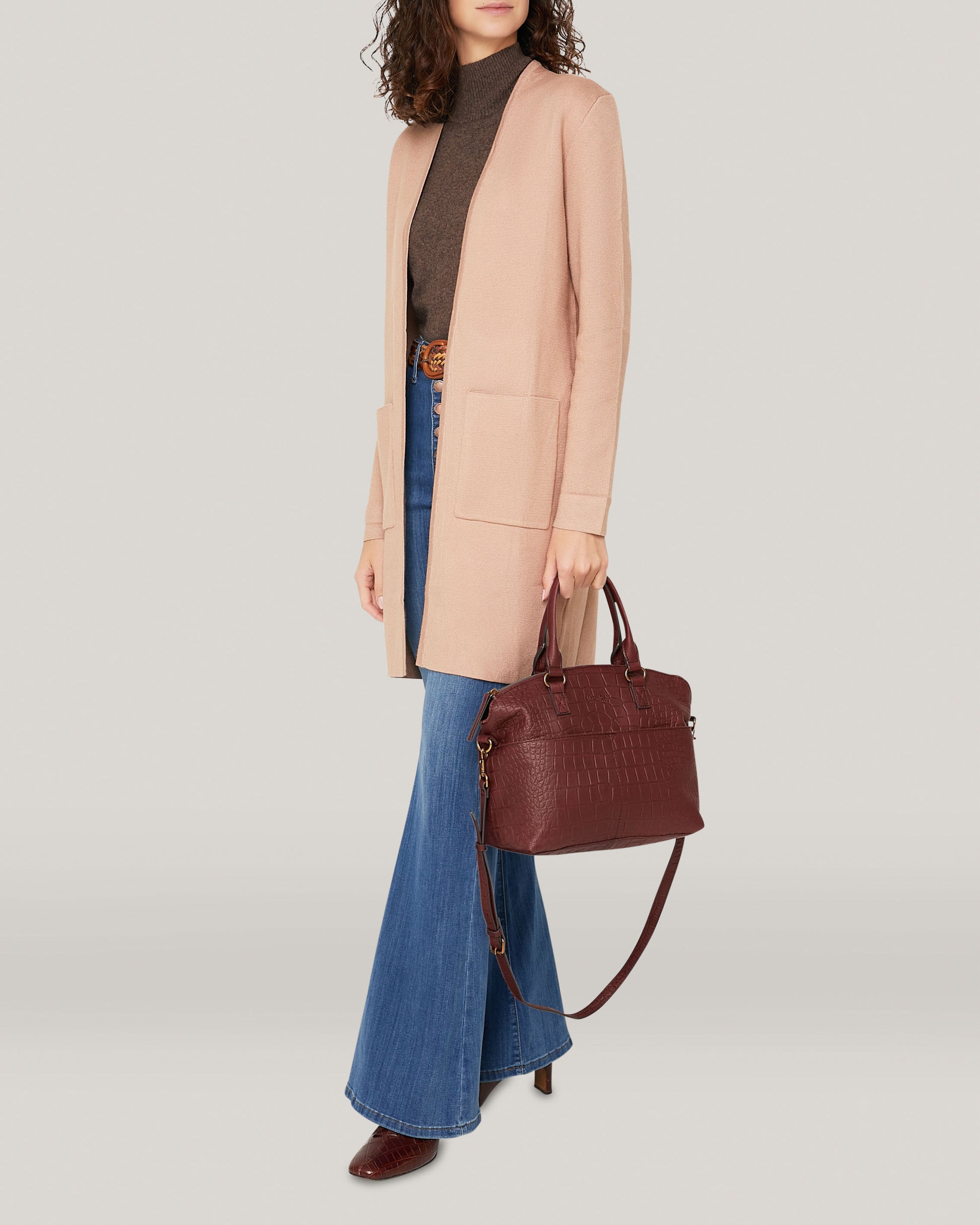 Wearing a long light pink coat, dark turtleneck sweater, and blue flared jeans, a person with curly hair holds an American Leather Co. Carrie Dome Satchel in burgundy with an adjustable crossbody strap against a plain, light background.