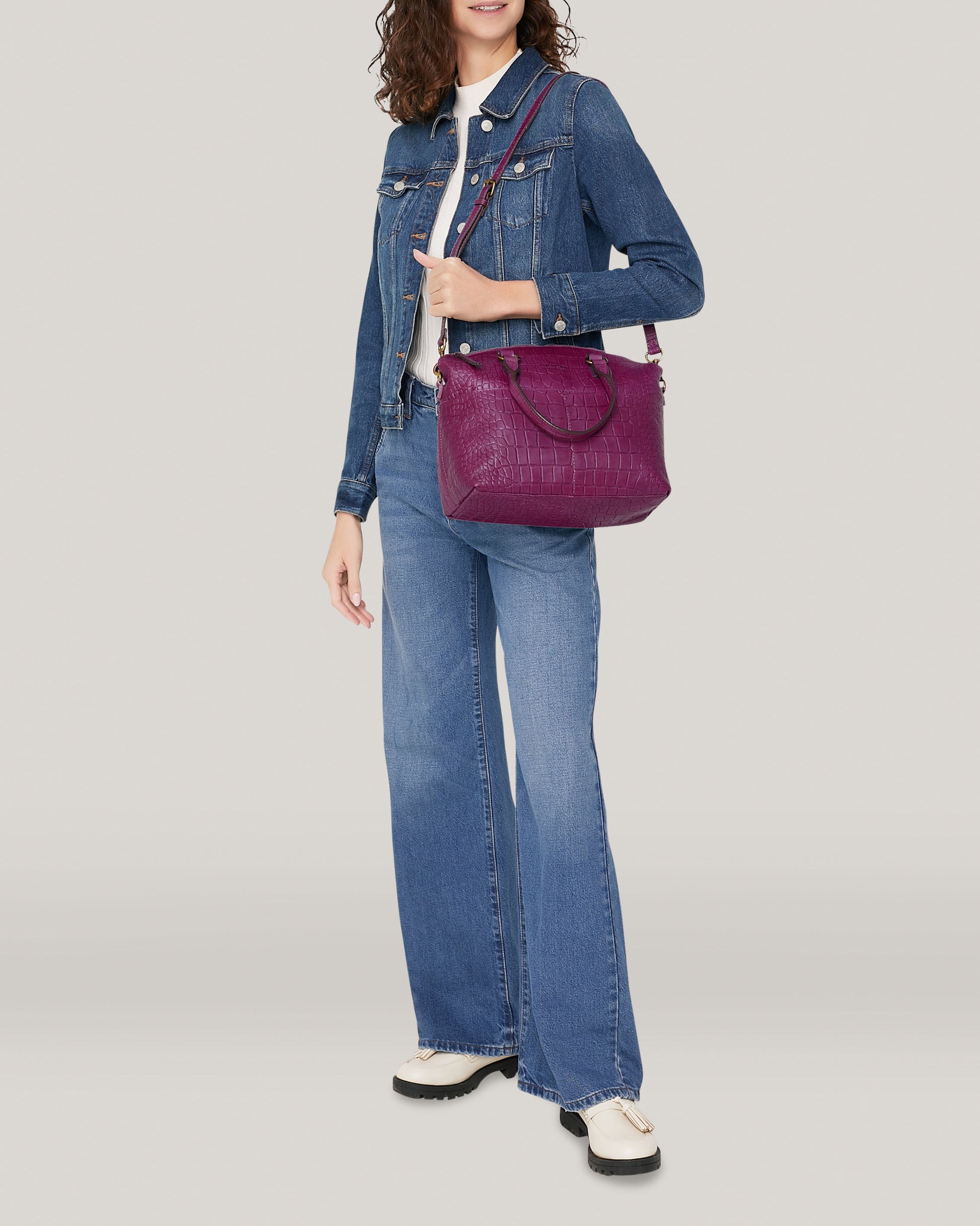 A person in a denim jacket and flared jeans holds an American Leather Co. Carrie Dome Satchel. They have curly hair and are wearing white shoes, with a plain off-white background behind them.