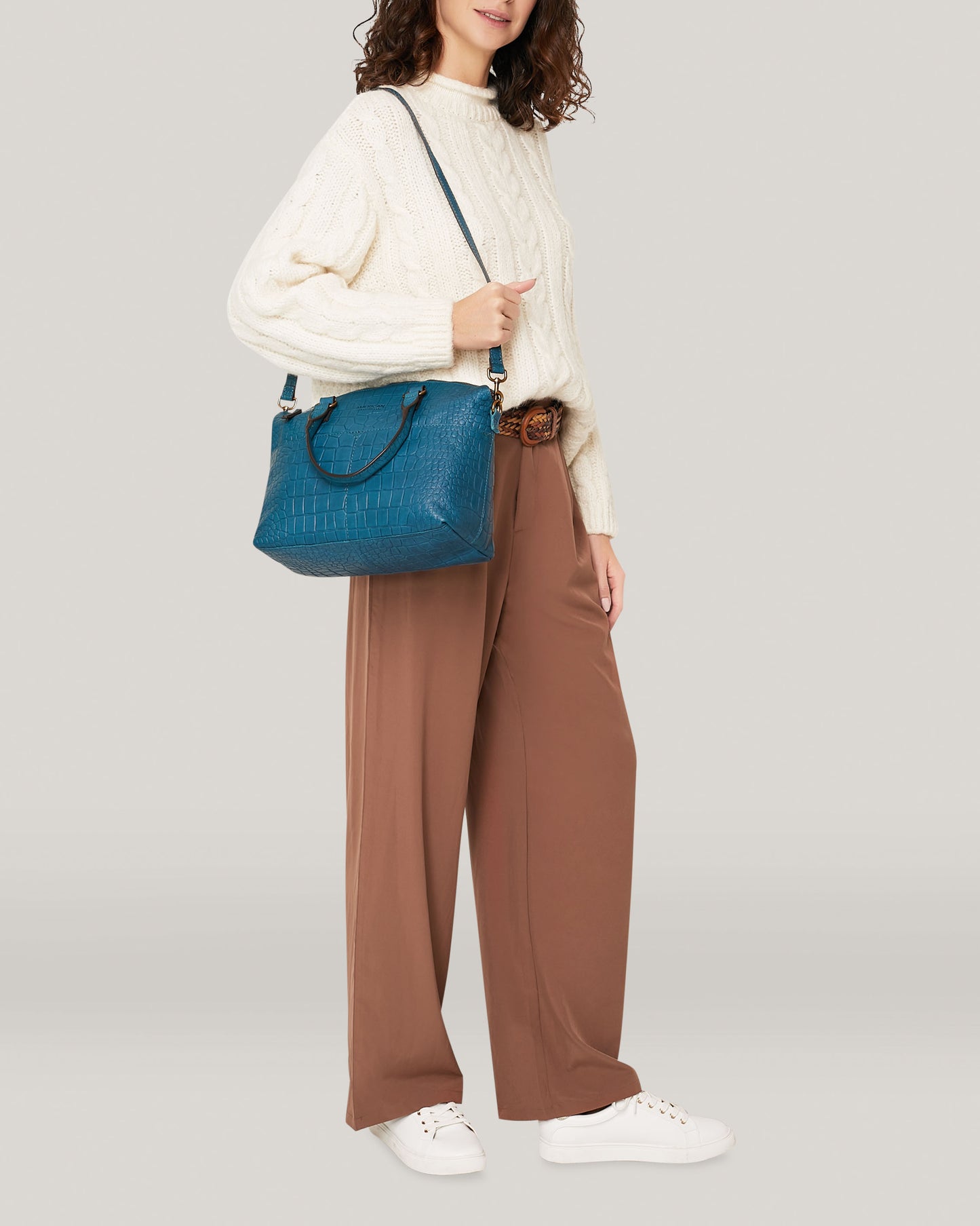 A person with curly hair is wearing a white cable-knit sweater, tan wide-leg pants, and white sneakers. They are carrying a blue Carrie Dome Satchel by American Leather Co., draped over their shoulder with its adjustable crossbody strap. The background is a solid light gray.